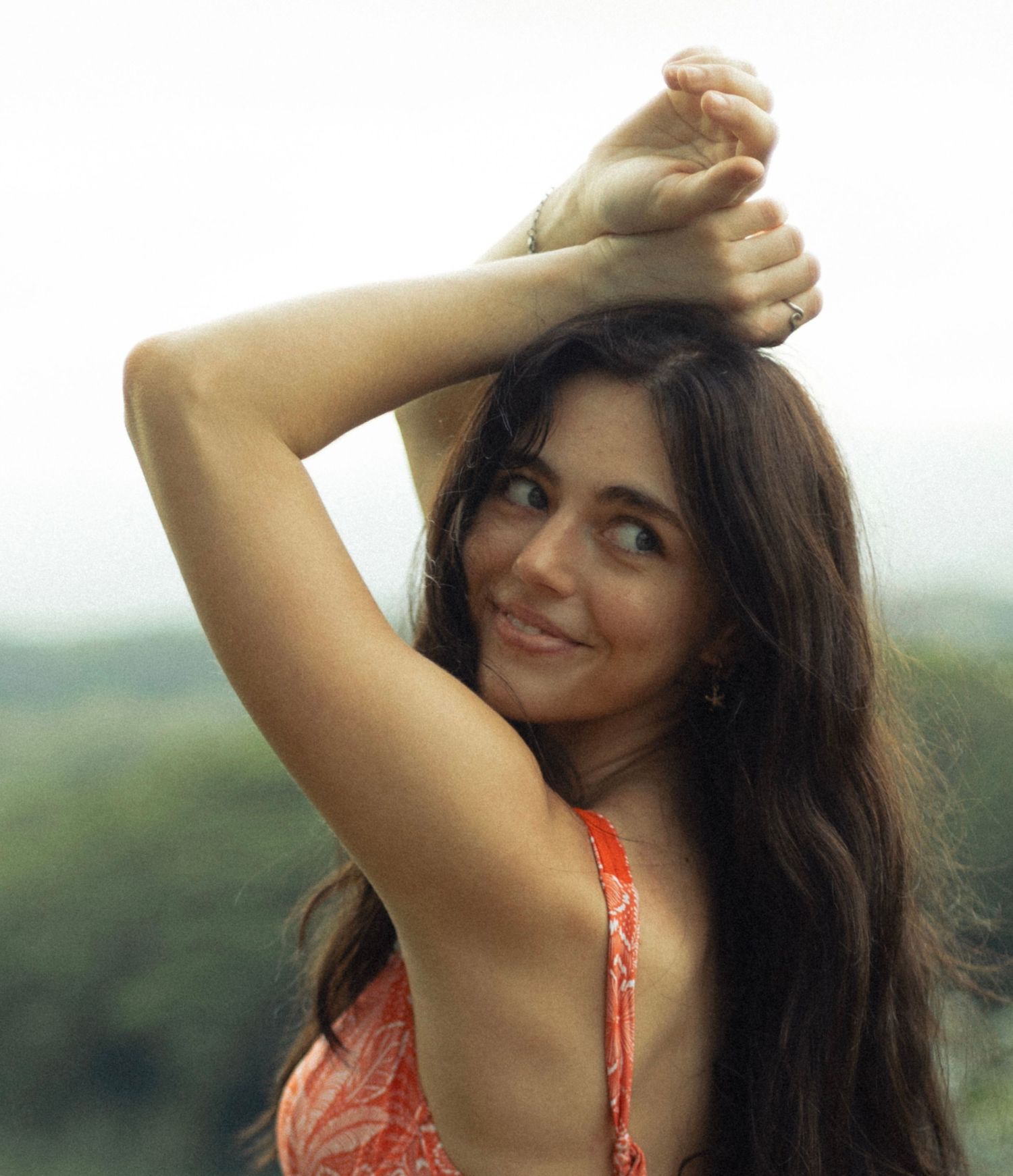 Woman in coral top poses with hands raised against blurred natural backdrop.