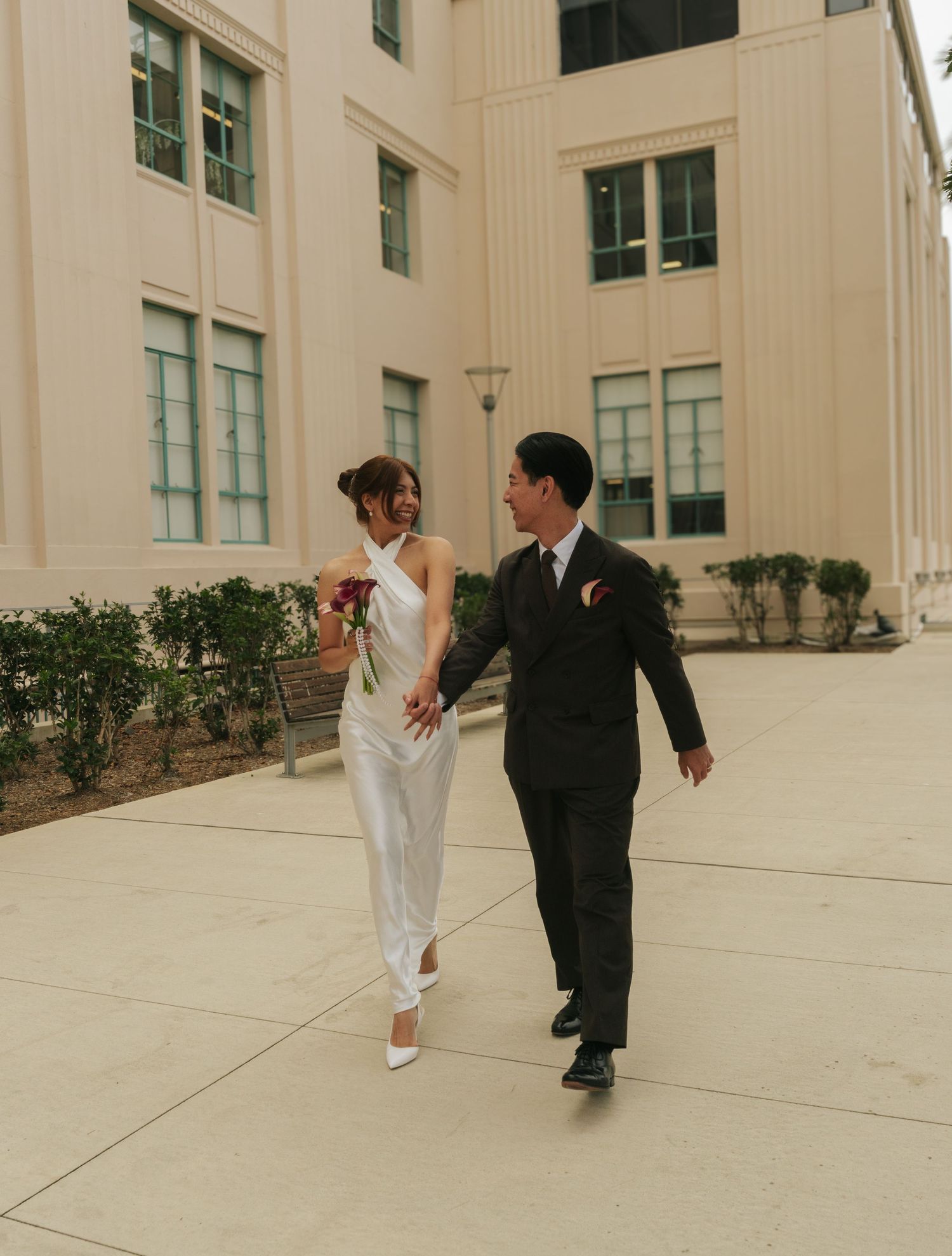 A bride in a white jumpsuit and groom in a black suit walk hand in hand outside a beige building.