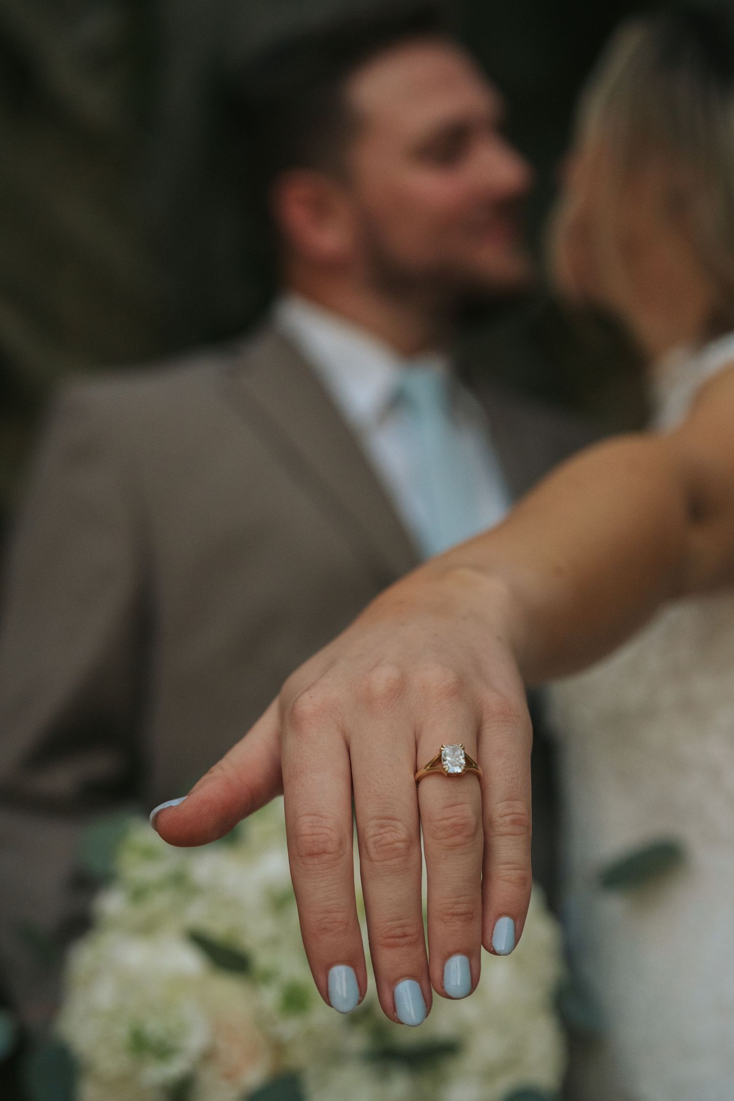 Close-up of bride's hand displaying elegant diamond engagement ring with couple kissing softly in background.