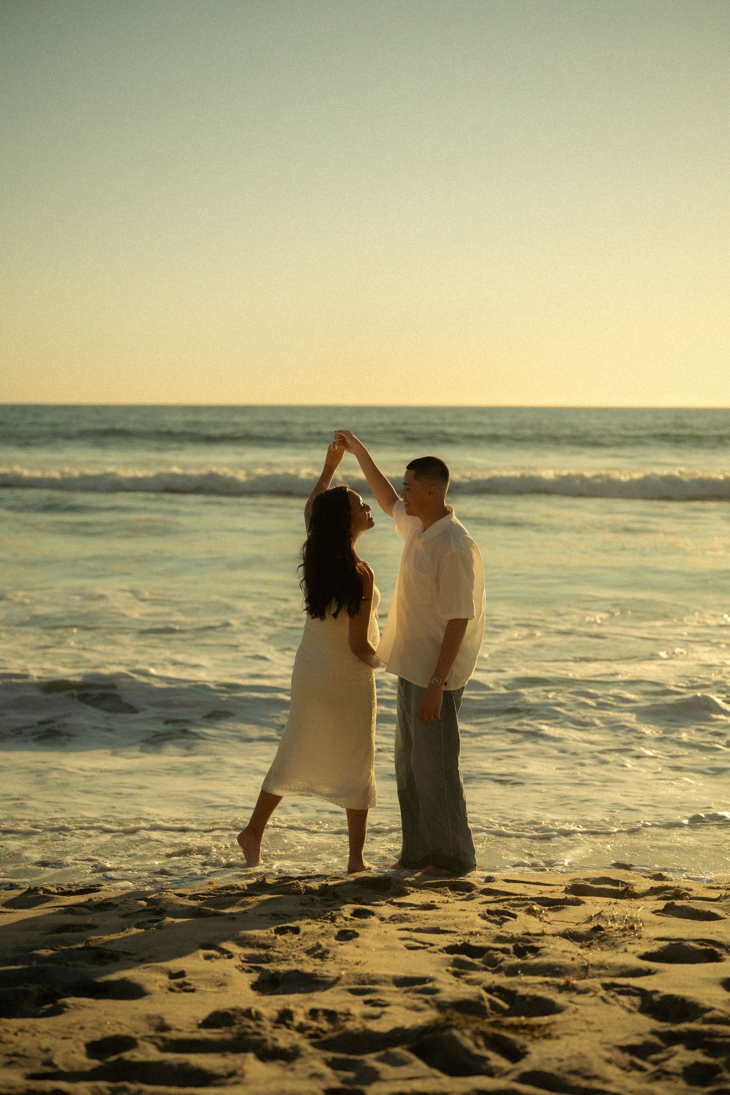 Couple dancing romantically on sandy beach at sunset, woman spinning gracefully with ocean waves behind them.