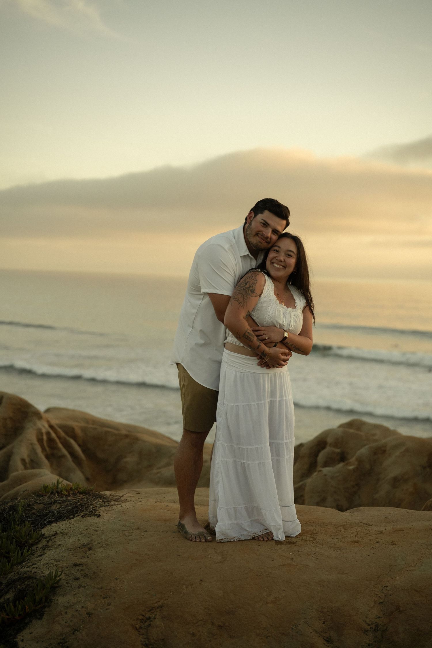 Couple embracing on rocky beach at sunset, woman in white dress, man in white shirt, ocean waves in background.