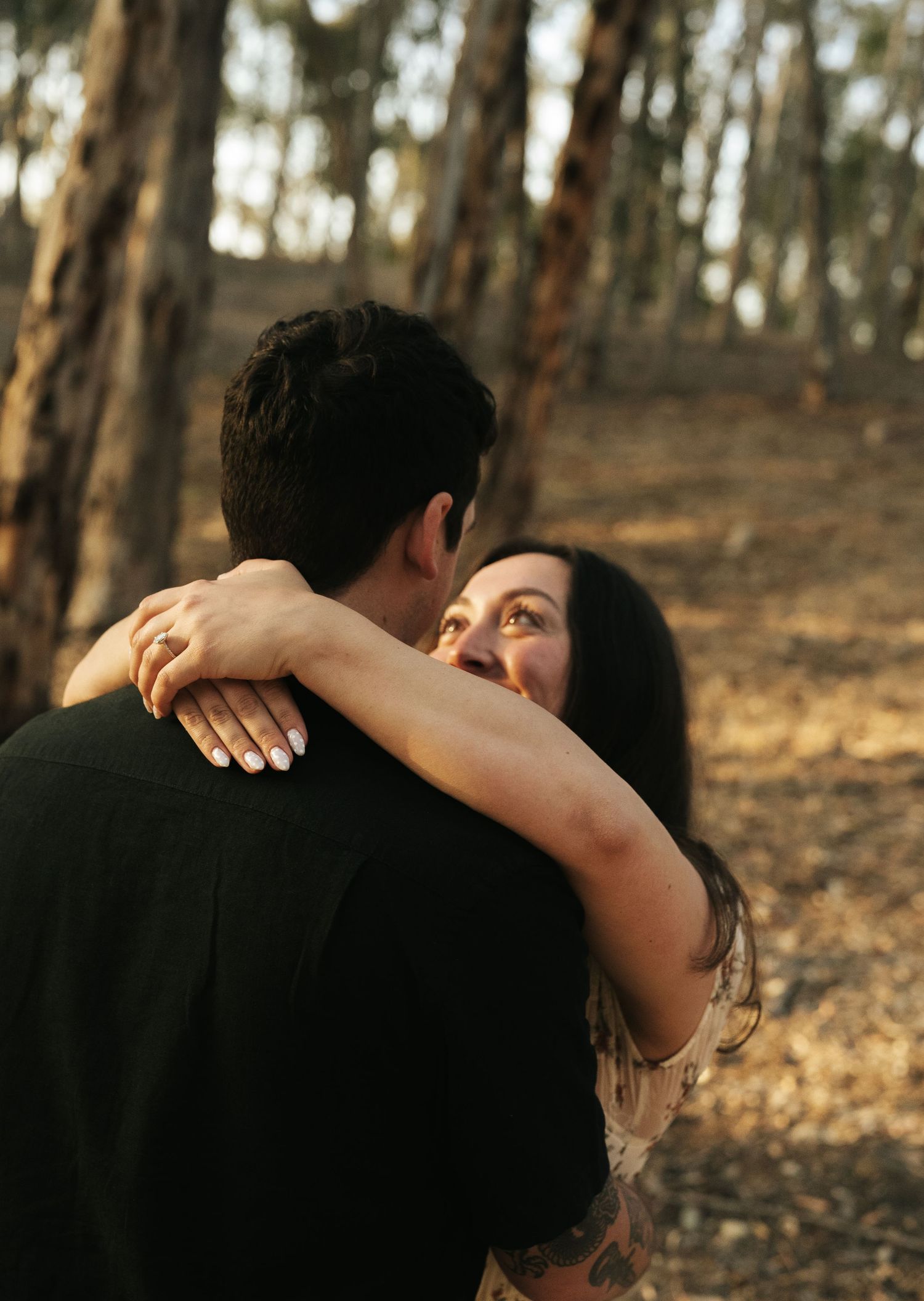 Woman with long dark hair embracing a man from behind in a sunlit pine forest, smiling softly.
