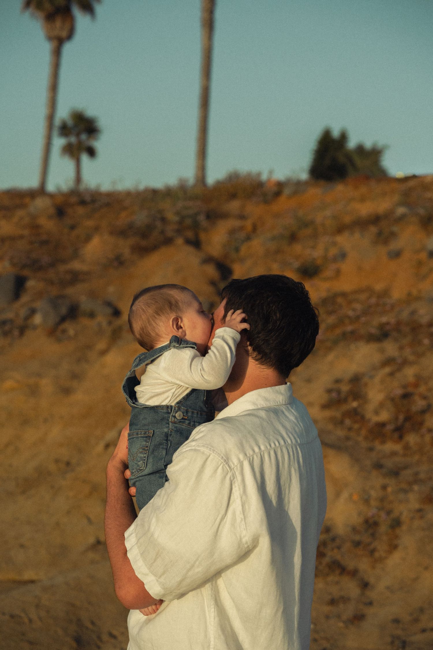A father tenderly holds his baby against a sandy hillside with palm trees at sunset.