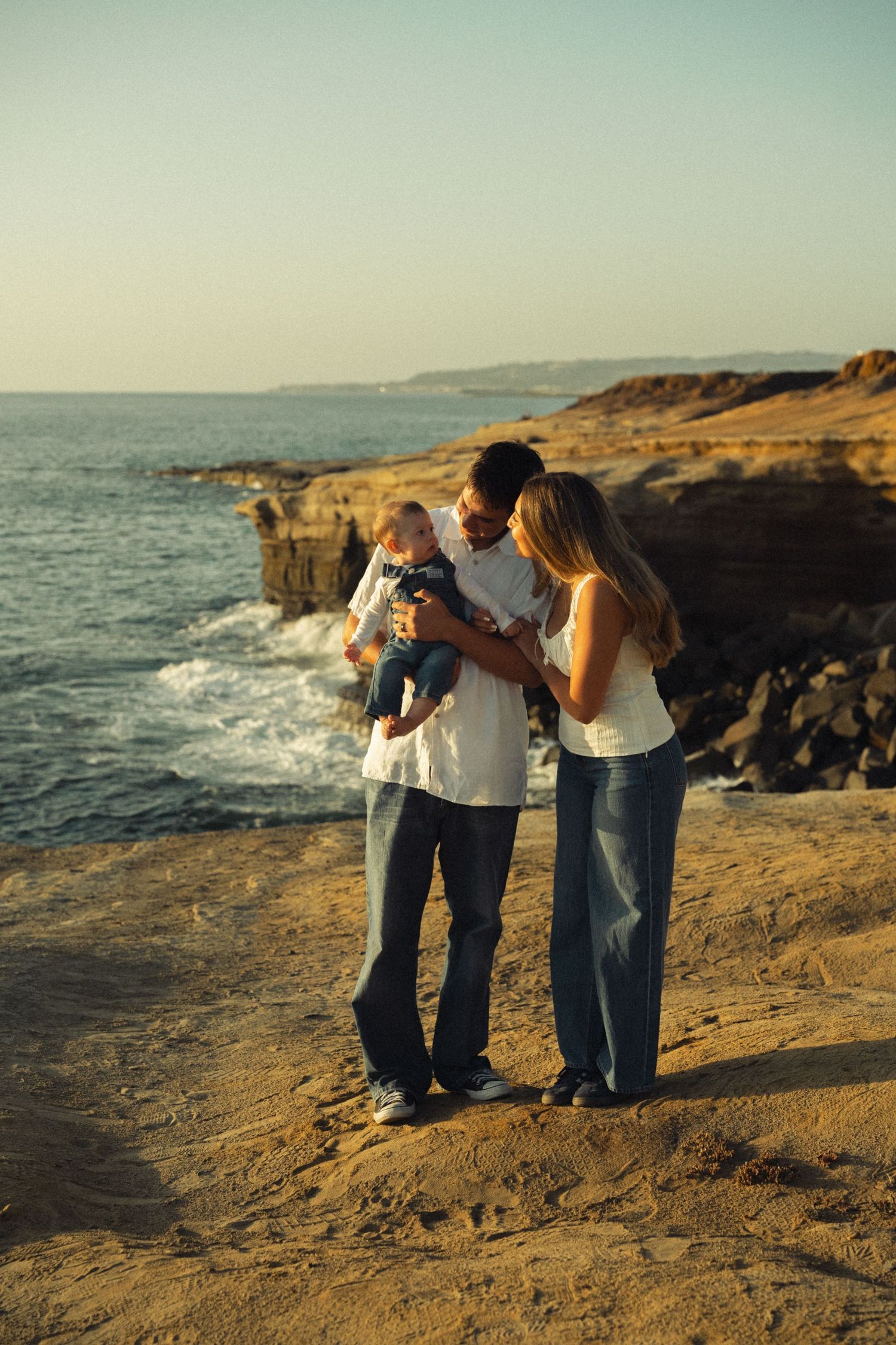 Family with infant standing on rocky ocean bluff during warm sunset photography session.