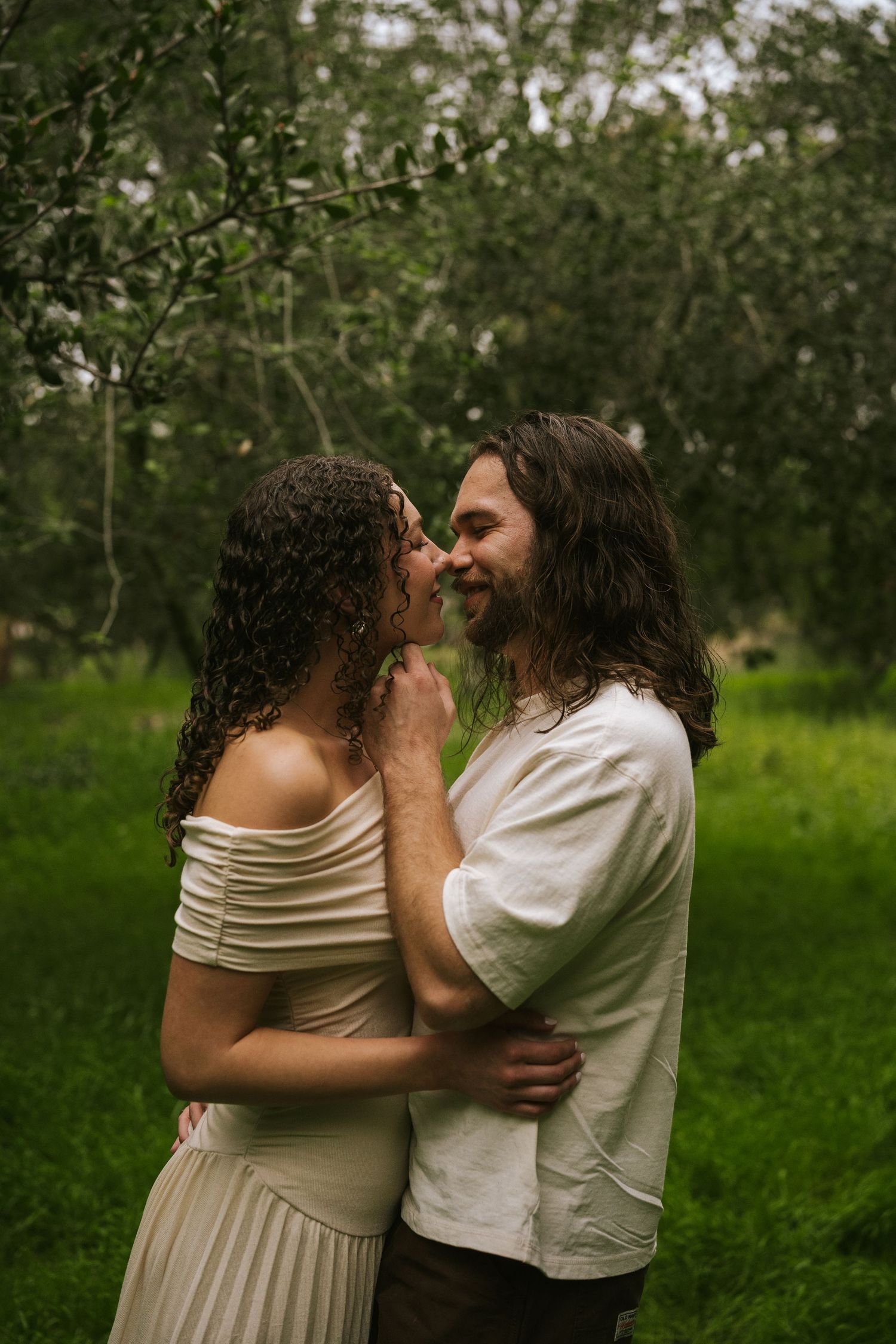A couple with long curly hair embraces and nearly kisses in a lush green garden, wearing cream and white outfits.