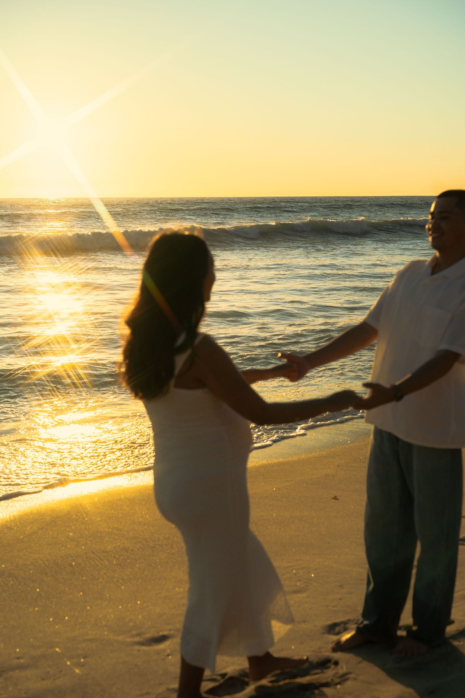 Couple holding hands on sandy beach at sunset with golden sun reflecting on ocean waves.