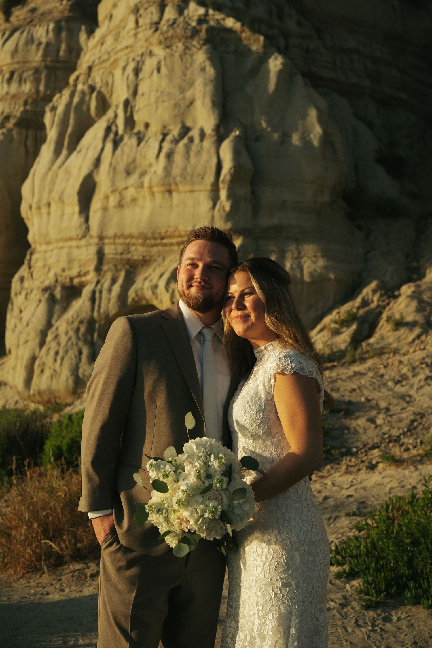 Bride and groom posing together in front of sandy cliffs at golden hour, bride holding white hydrangea bouquet.