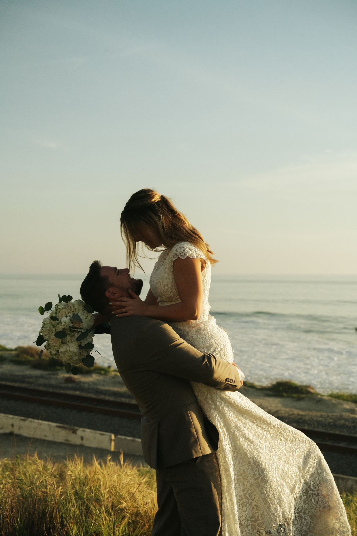 Groom lifts bride at sunset beach wedding, holding white bouquet as waves crash behind the couple.