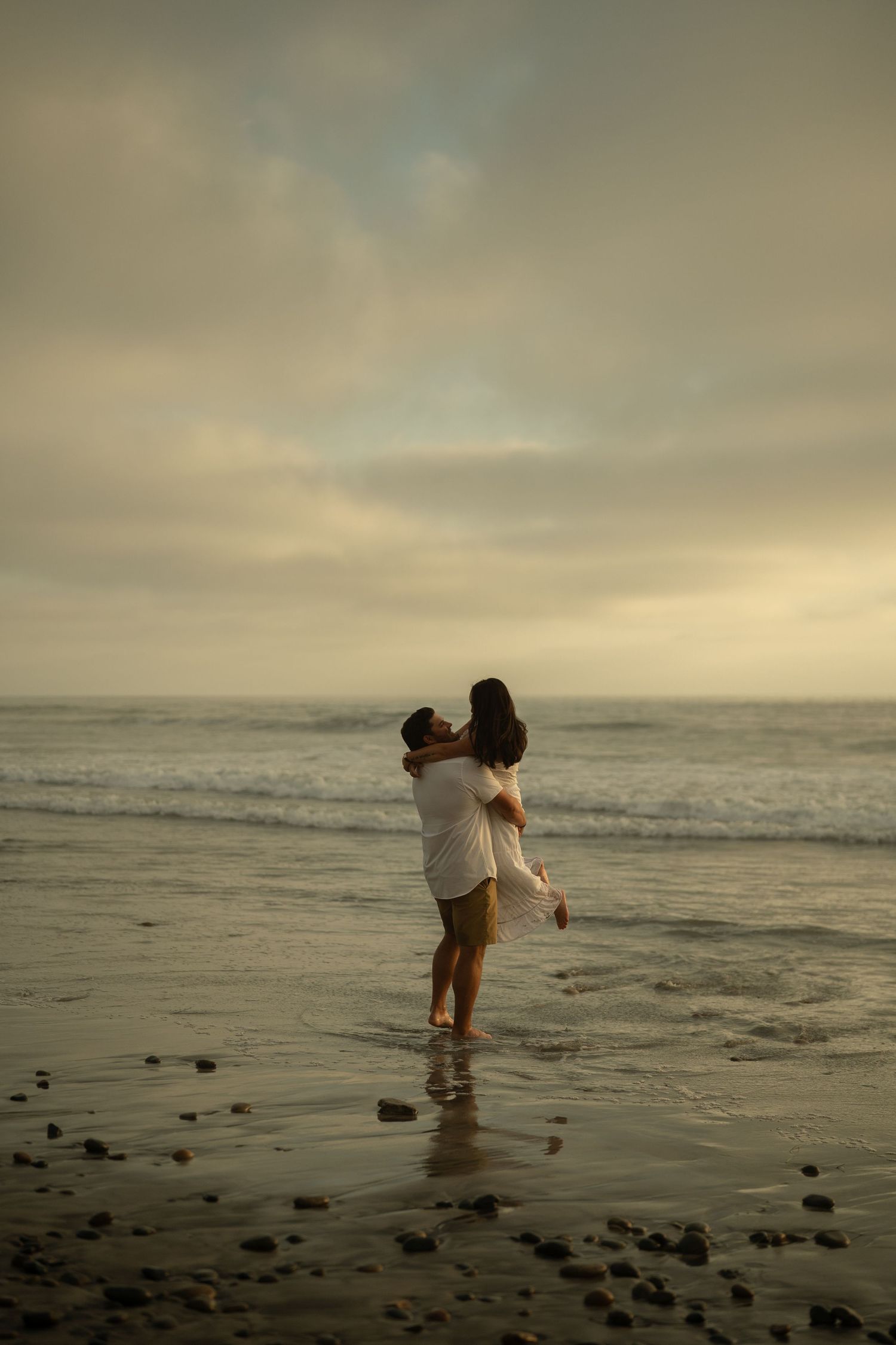 Couple embracing romantically on a misty beach at sunset, with ocean waves and cloudy sky in the background.