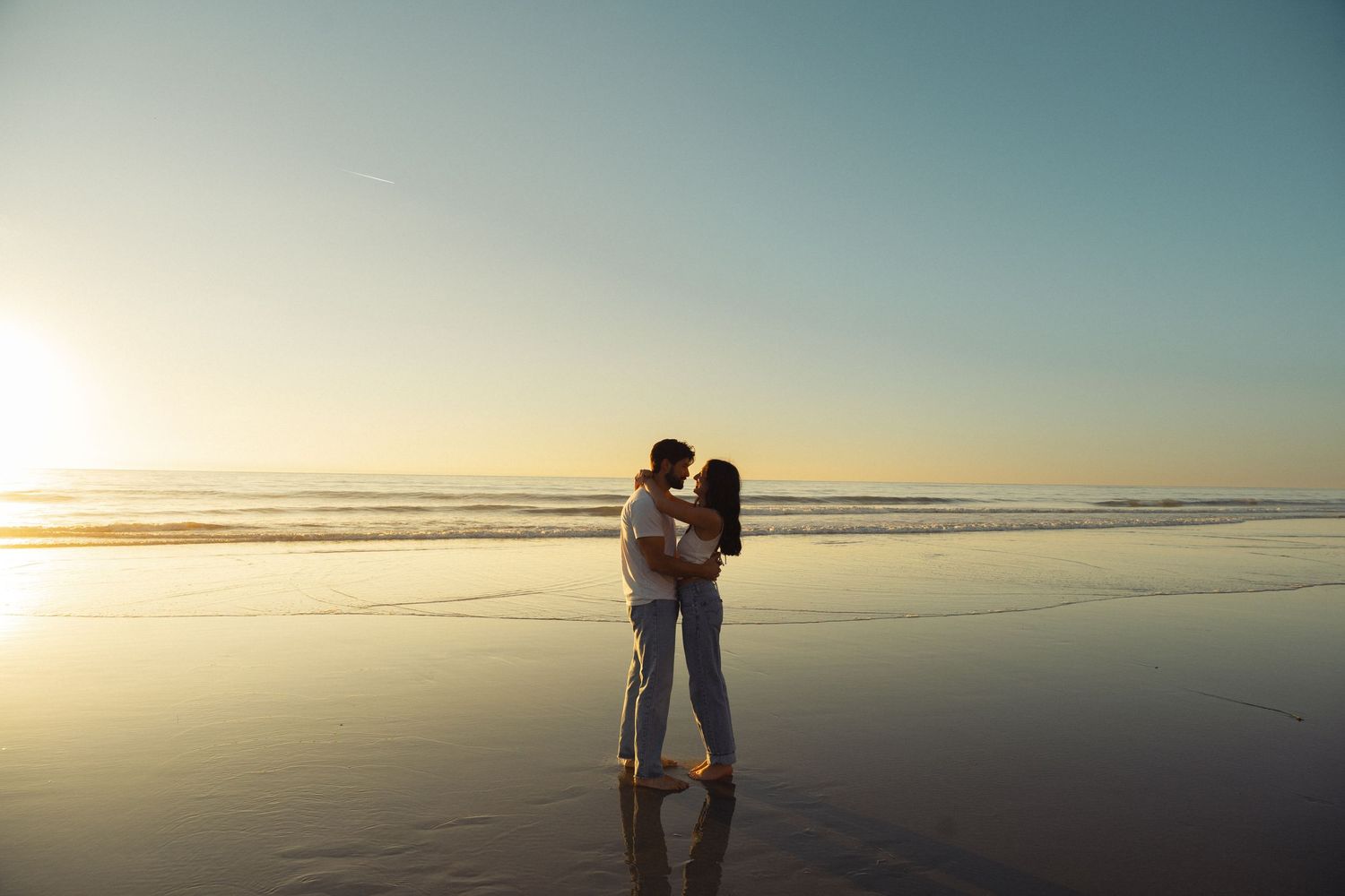 Romantic couple embraces on a beach at sunset with gentle waves in the background.
