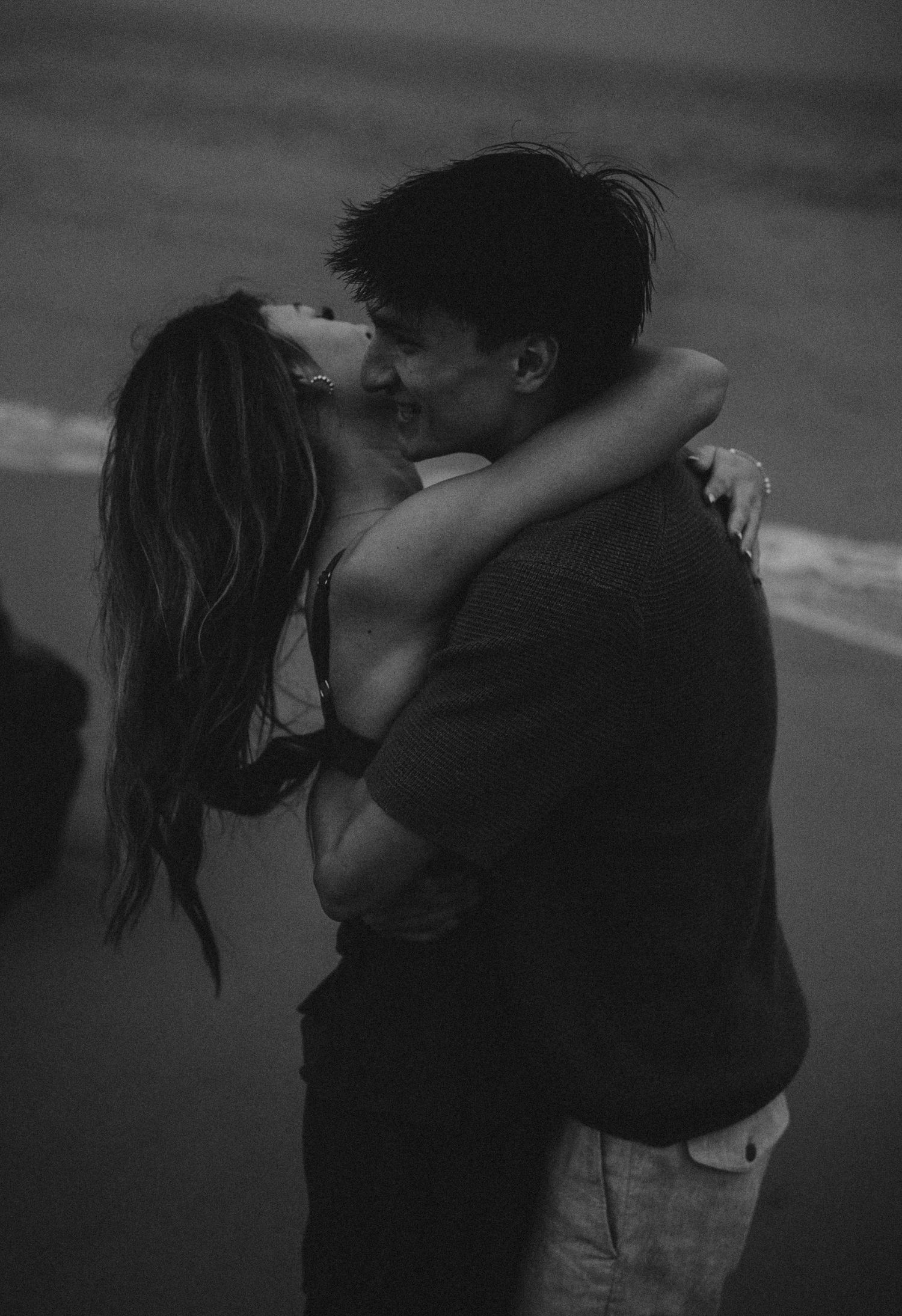 Black and white photo of a romantic couple embracing and kissing on a beach at dusk.