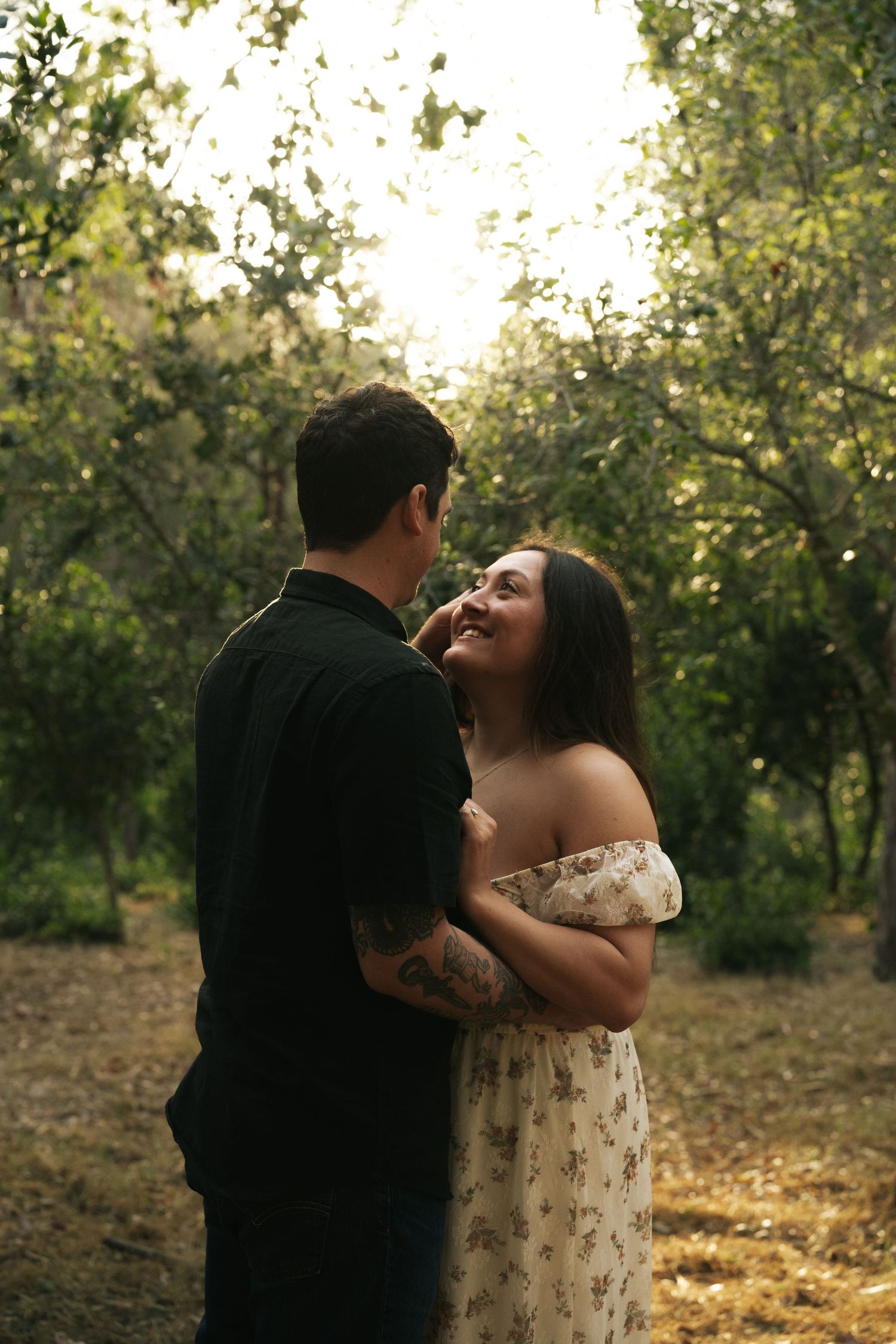 Couple sharing an intimate moment in a glowing forest, woman in floral dress laughing up at her partner.