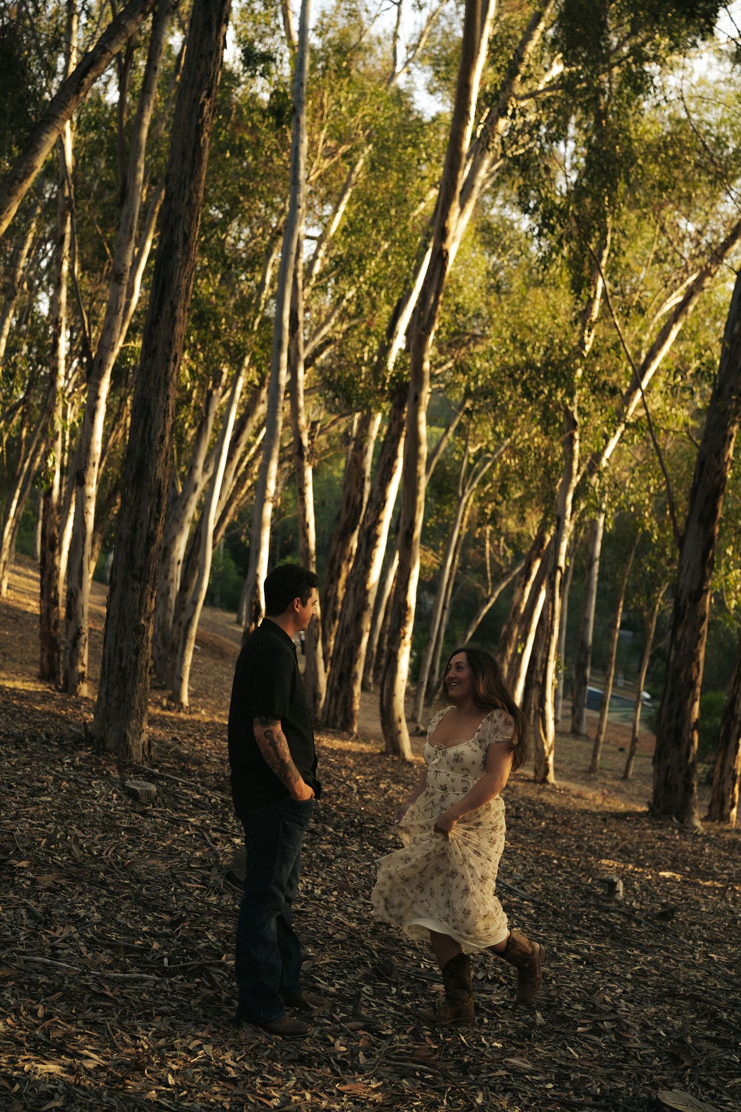 Couple holding hands and dancing playfully among tall trees in golden hour sunlight in a woodland setting.