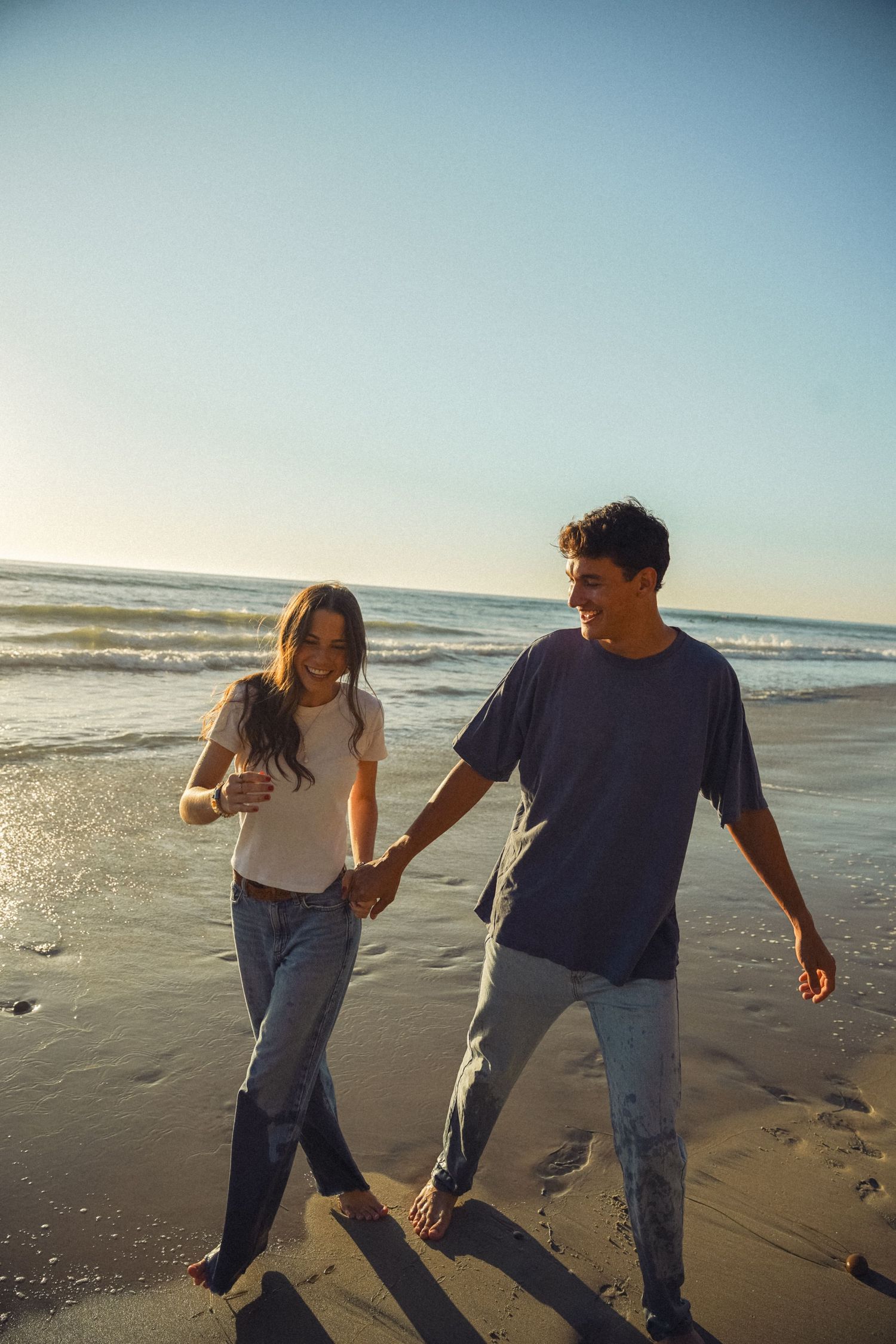A happy young couple holds hands while walking barefoot on a sunny beach with ocean waves in the background.