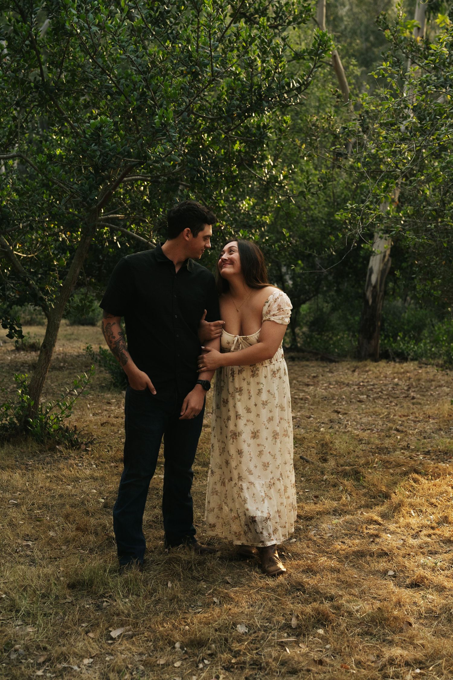 A joyful couple laughs together in a sunlit orchard, woman in floral dress embracing man in black shirt.
