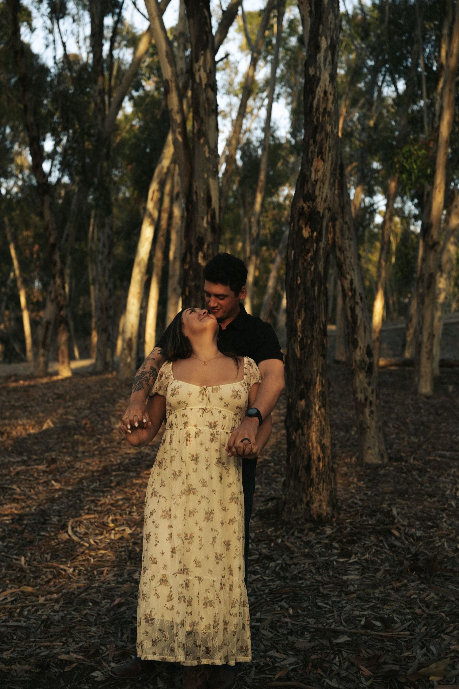 A couple shares a tender kiss in a sunlit forest, the woman wearing a floral dress holding hands with her partner.