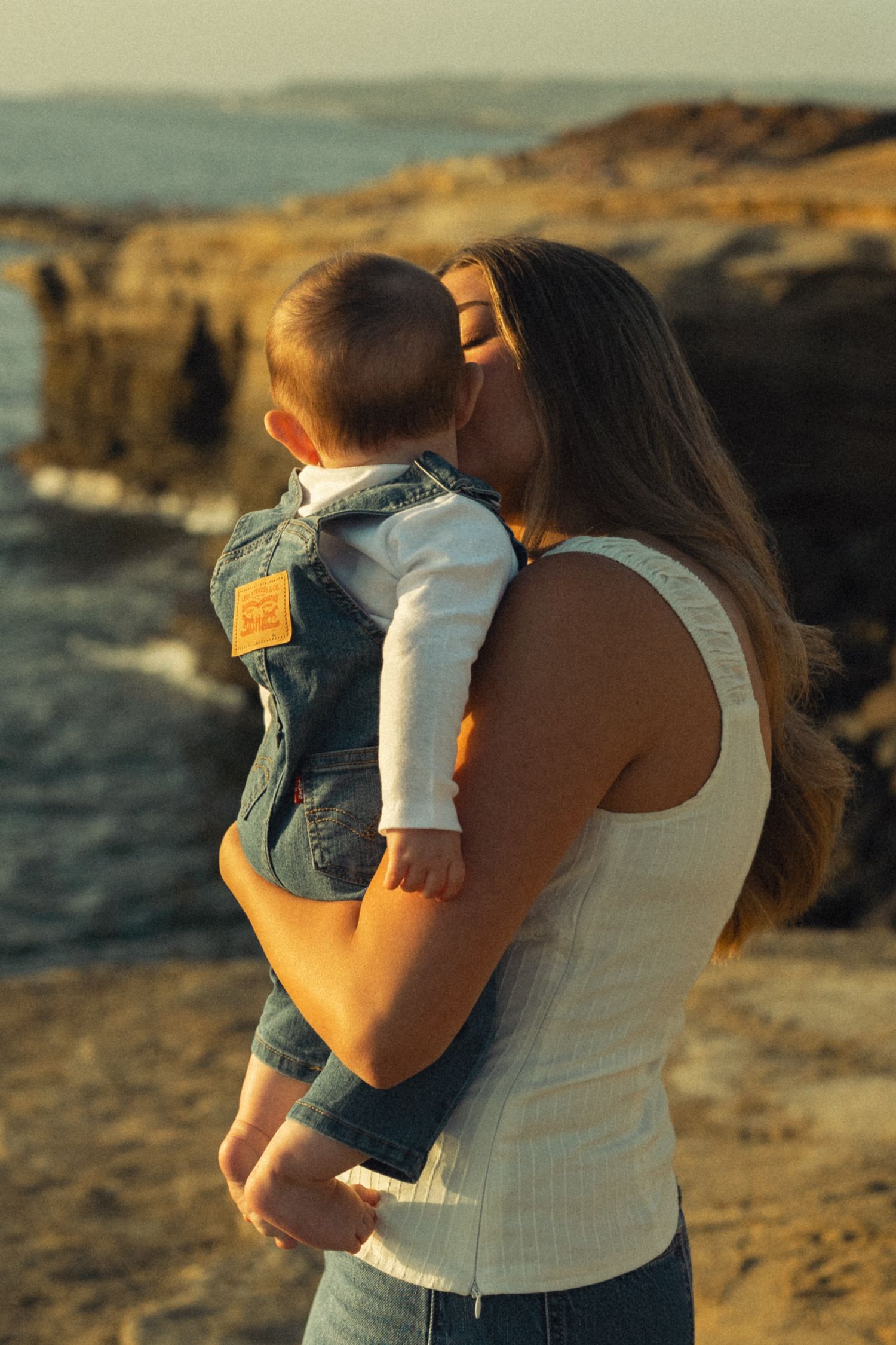 A mother tenderly kisses her baby outdoors near rocky coastal cliffs during golden hour sunset.