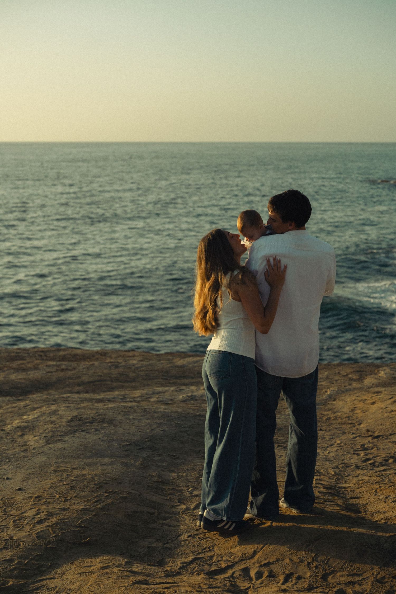 Family of three standing together embracing while gazing at calm ocean horizon at dusk.