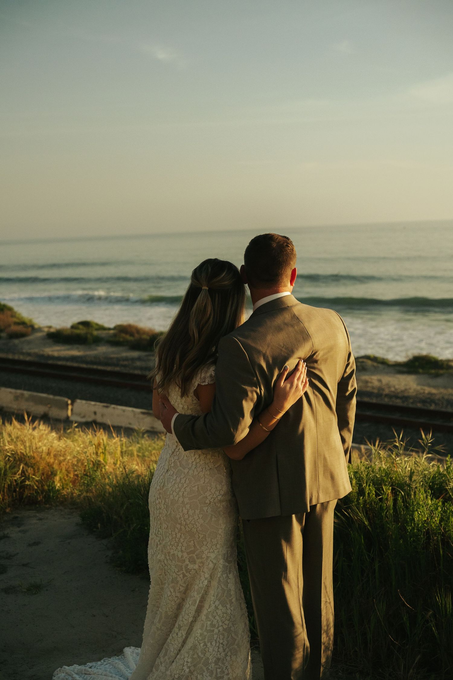 Newlywed couple embracing while gazing at ocean waves during golden hour beach wedding portrait.