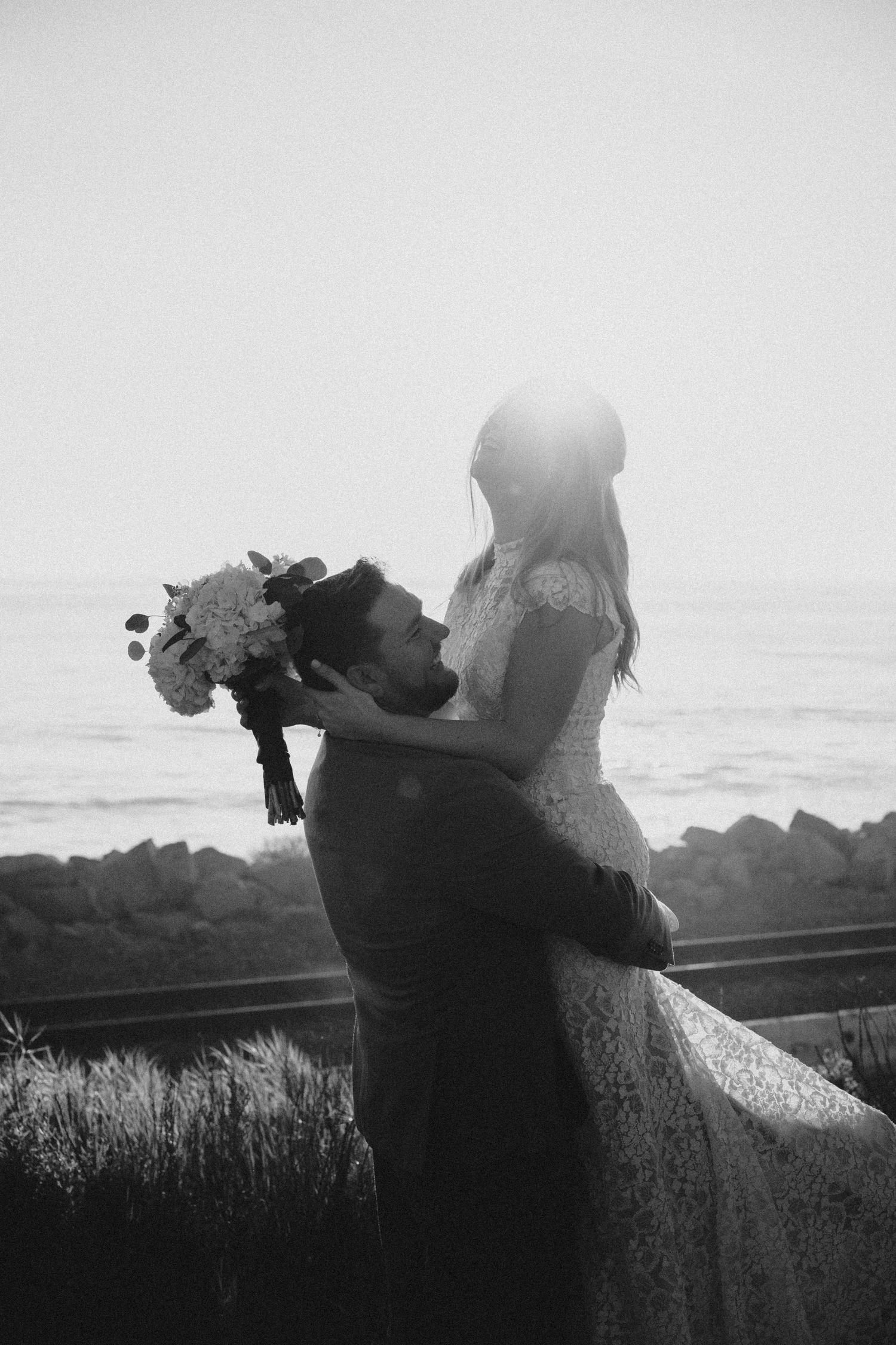 Black and white photo of groom lifting bride by the ocean, bride holding a white floral bouquet.