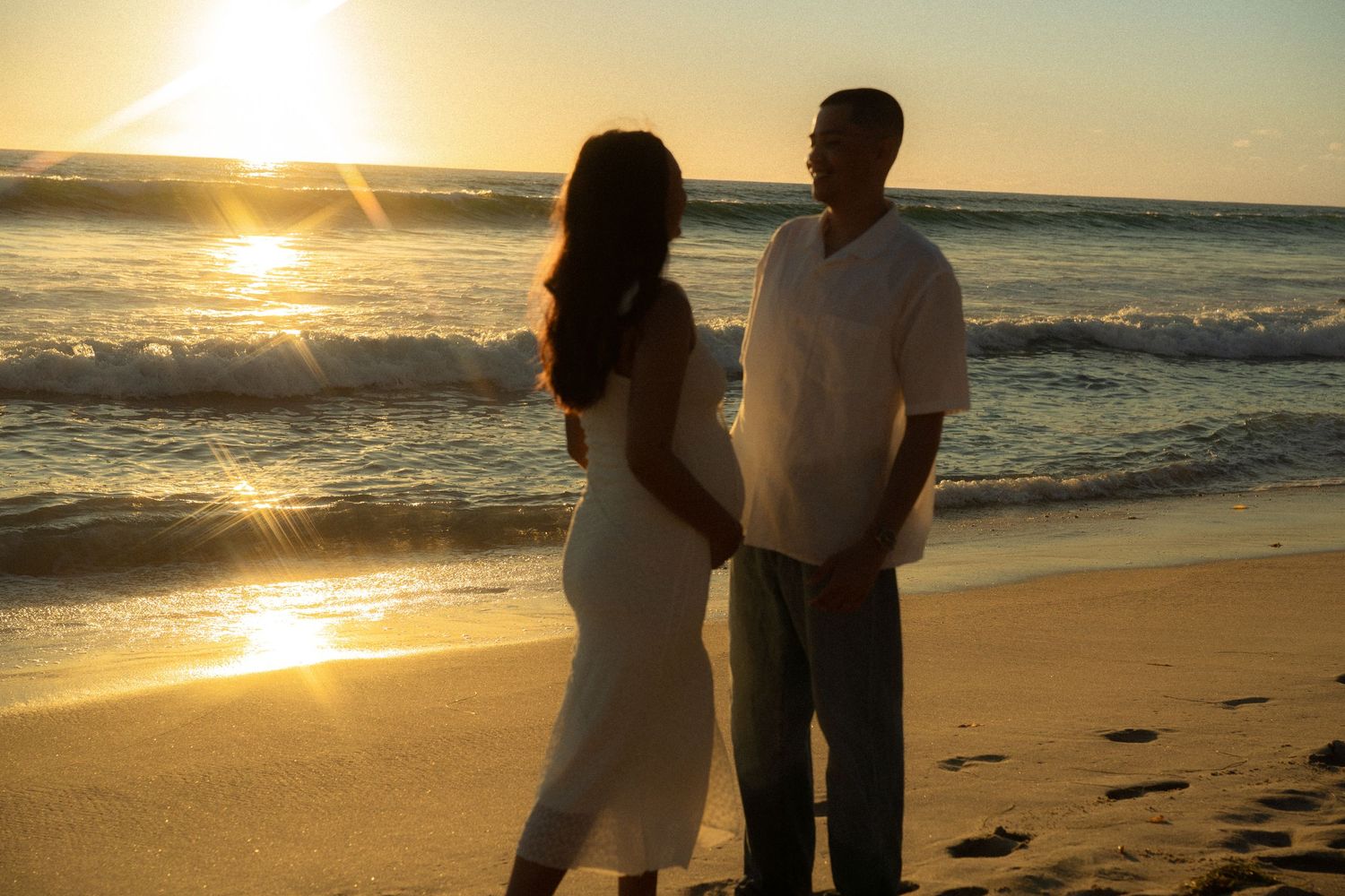 Couple holding hands on a sandy beach at sunset with golden light reflecting on ocean waves.