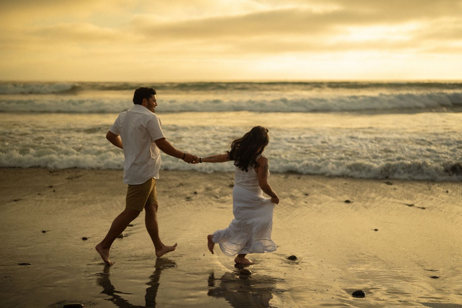 Couple holding hands running on a sandy beach at sunset, wearing white outfits with ocean waves in the background.
