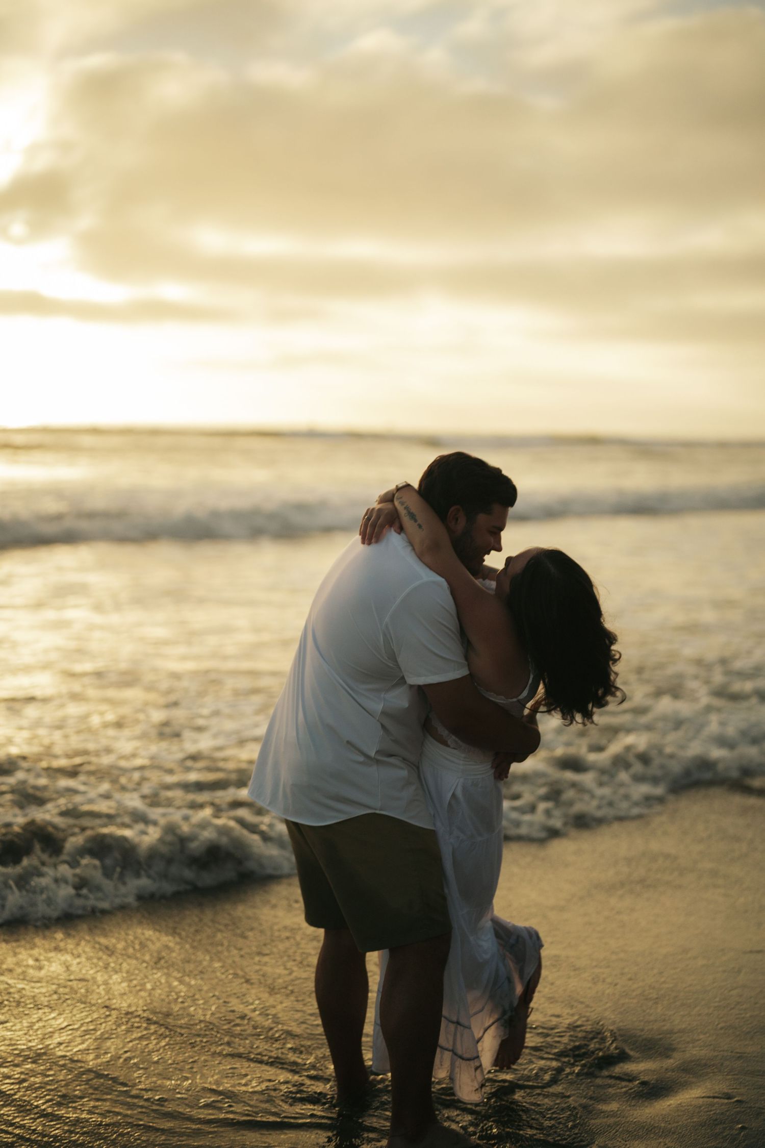 A couple shares a romantic kiss while standing in ocean waves at sunset on a beautiful beach.
