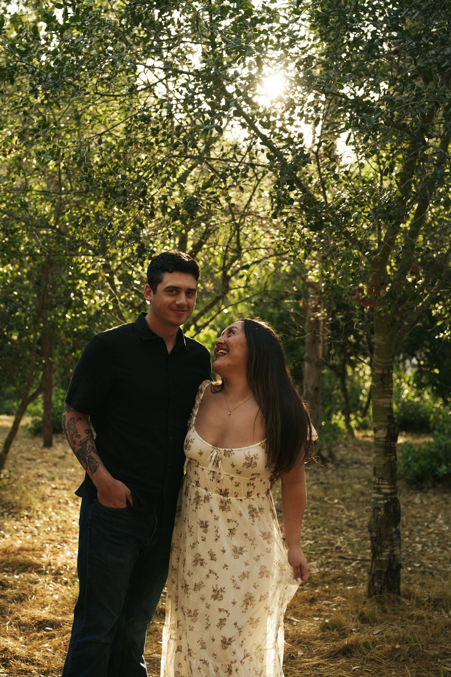 Couple laughing and walking together through sun-dappled trees during golden hour outdoor photoshoot.