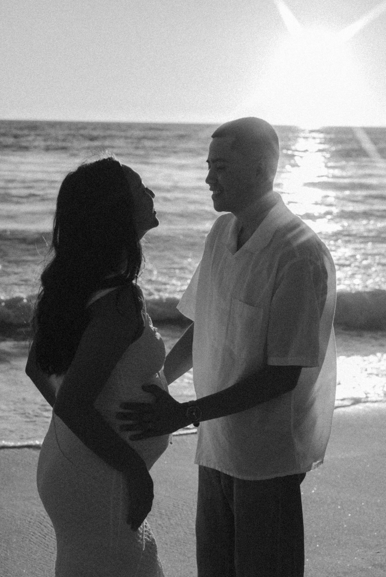 Black and white photo of pregnant couple facing each other on beach with sunlit ocean backdrop.