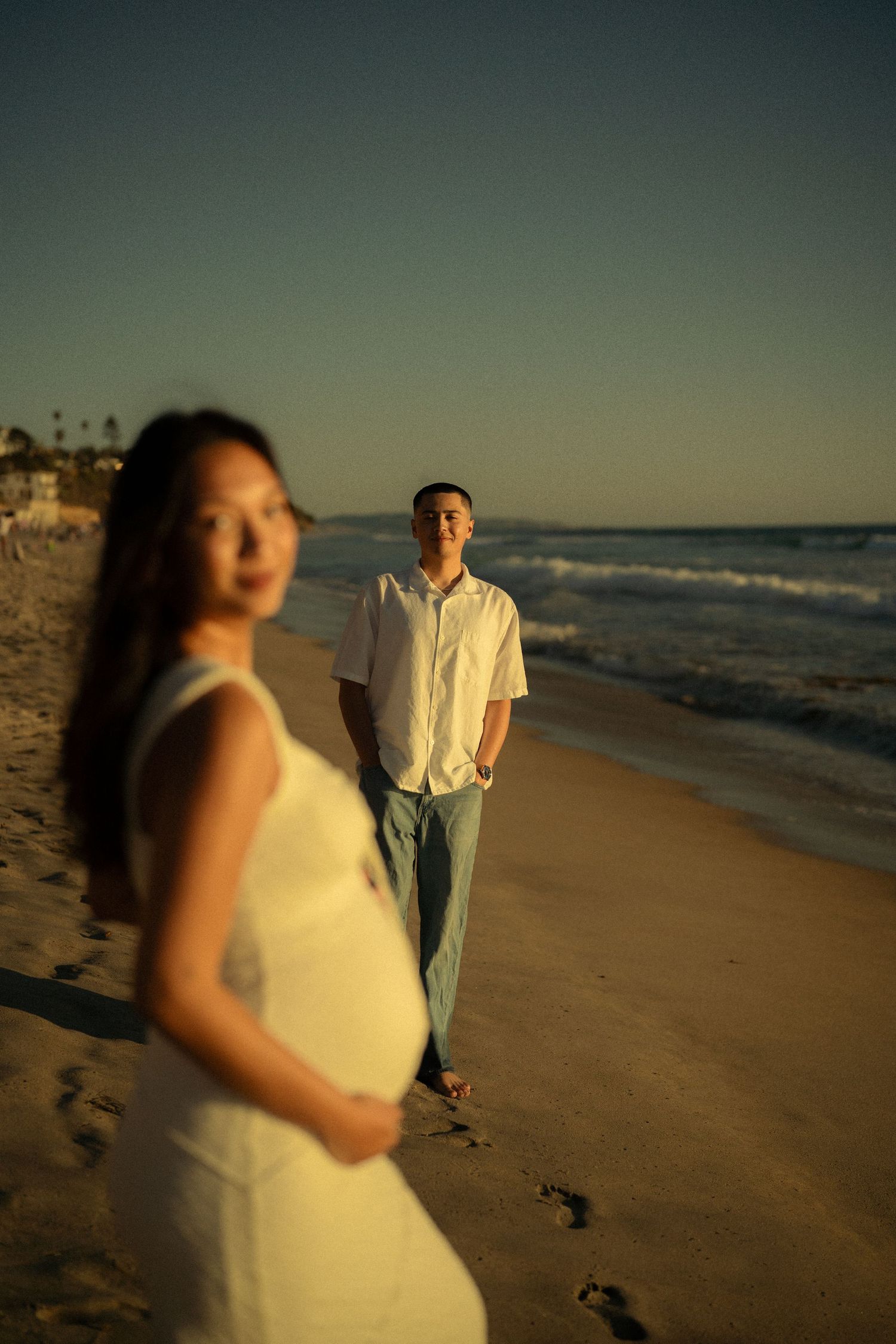 Pregnant woman in white dress with man in white shirt standing on beach at golden sunset.