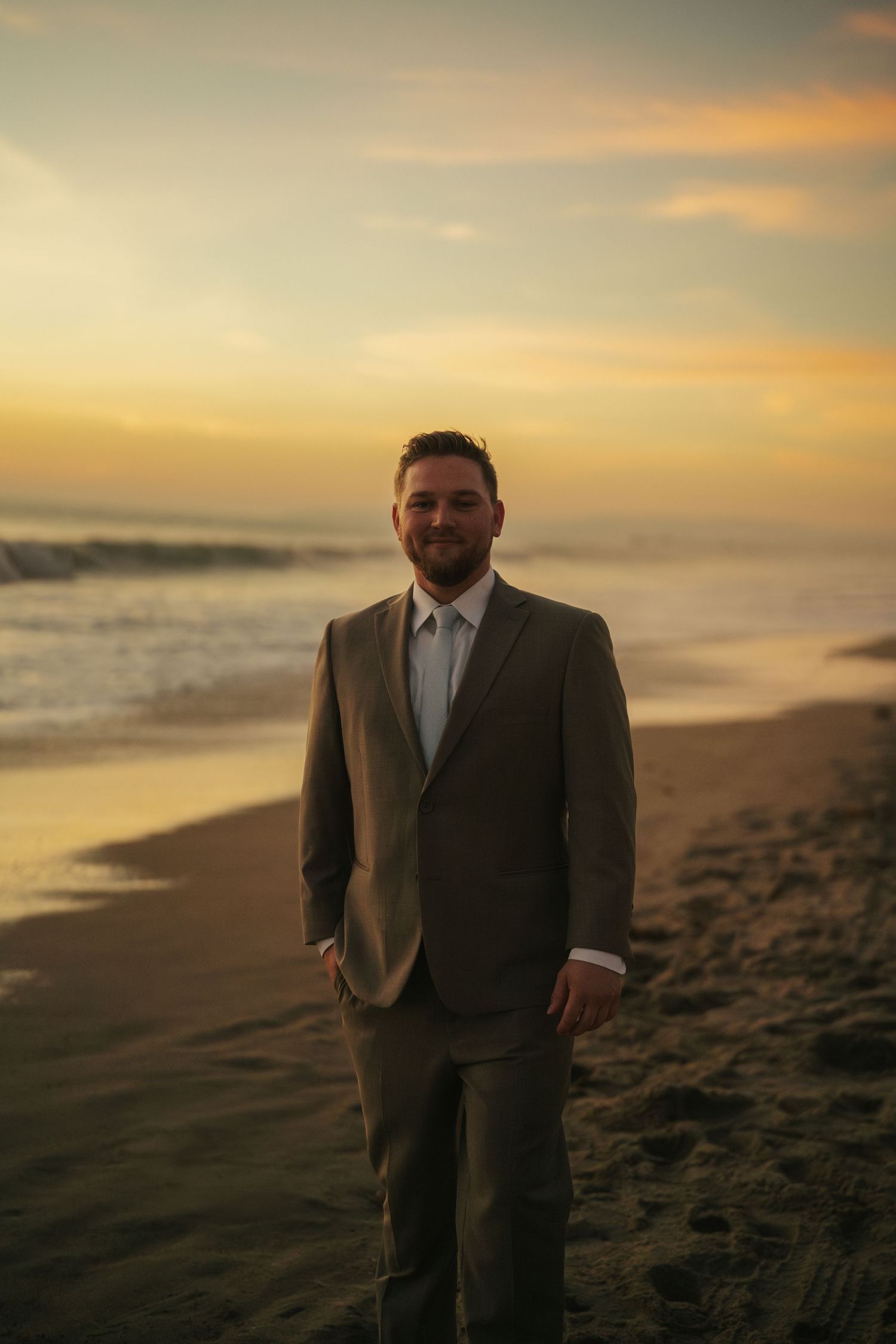 Handsome groom in tan suit stands confidently on sandy beach during vibrant golden sunset.