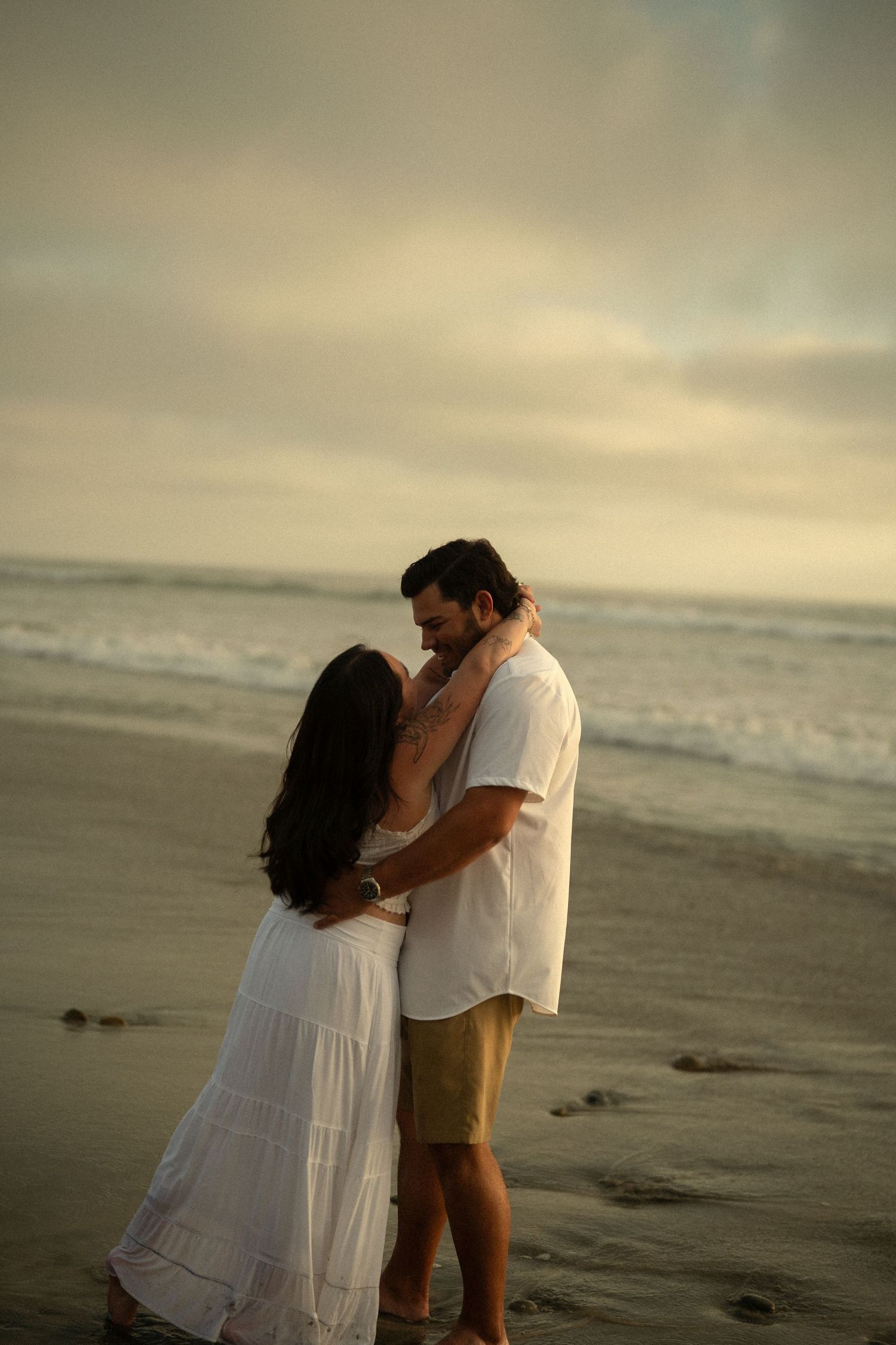 A loving couple shares an intimate moment on a sandy beach at sunset, woman in white dress embracing man in white shirt.