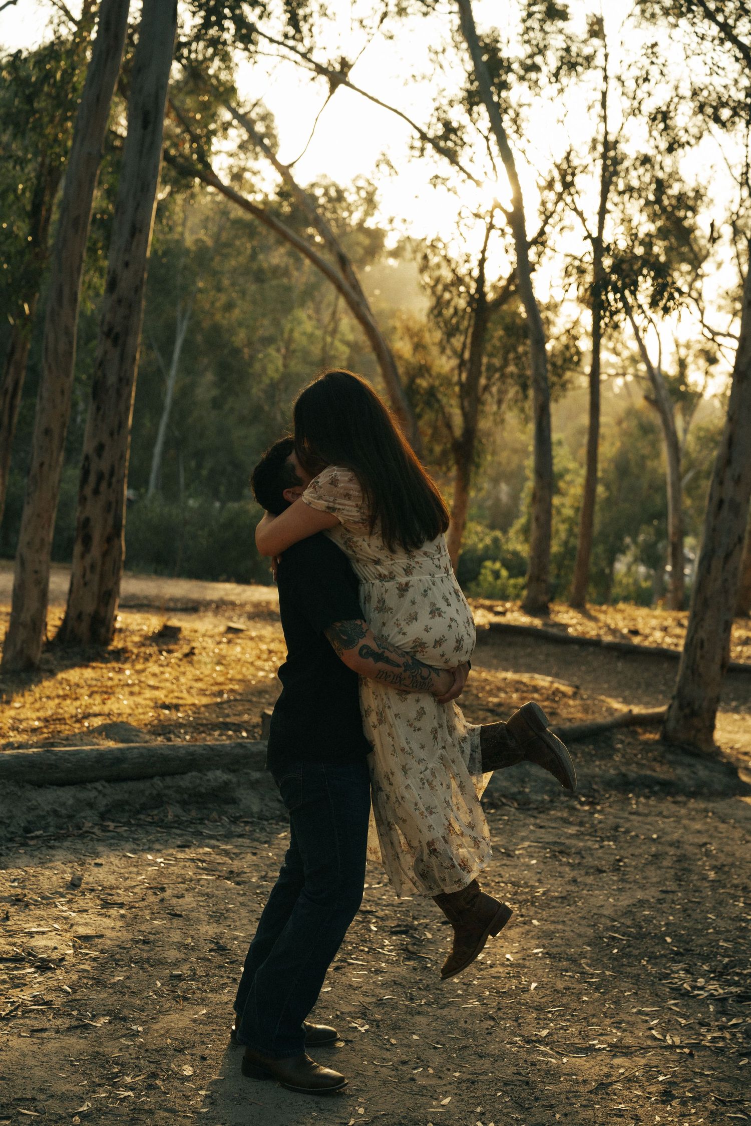 Man lifting woman in floral dress in a warm golden sunlit forest clearing.