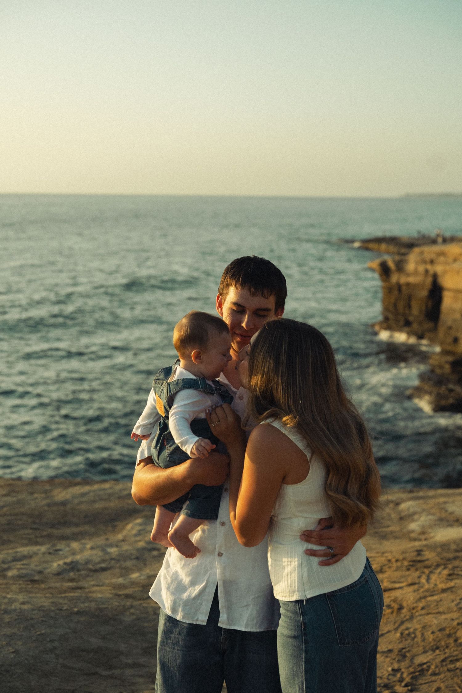Loving couple sharing a kiss over their baby while standing on rocky ocean cliffside at sunset.