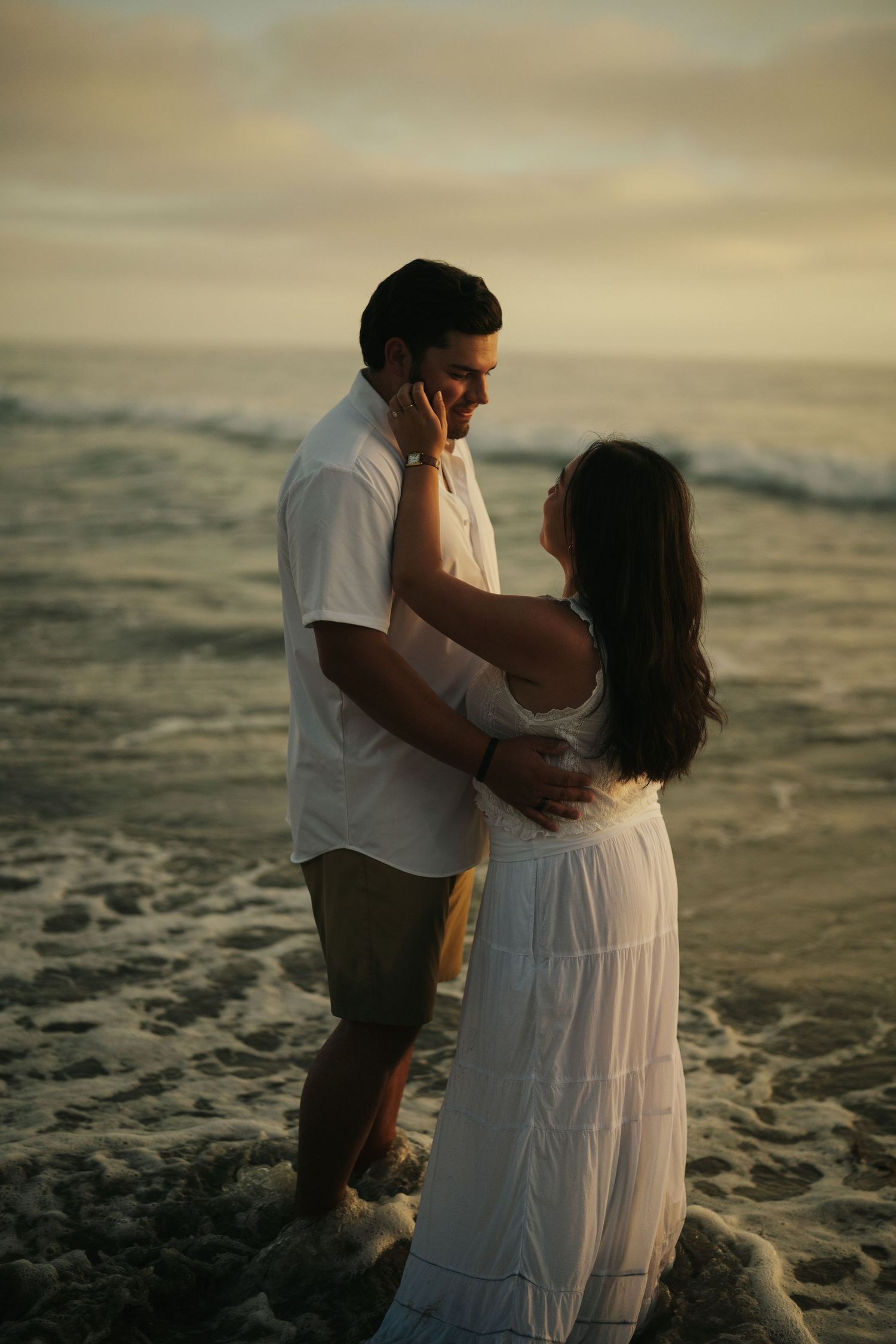 Romantic couple embracing in ocean waves at sunset, woman touching mans face in a tender moment.