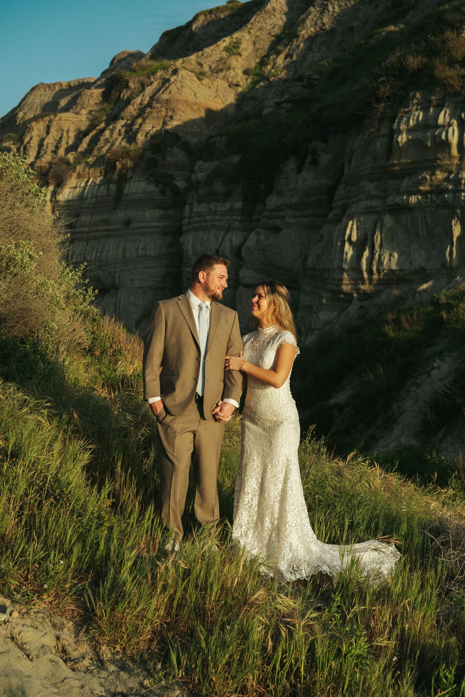 Wedding couple smiling together on grassy hillside with dramatic sandy cliffs backdrop in golden light.