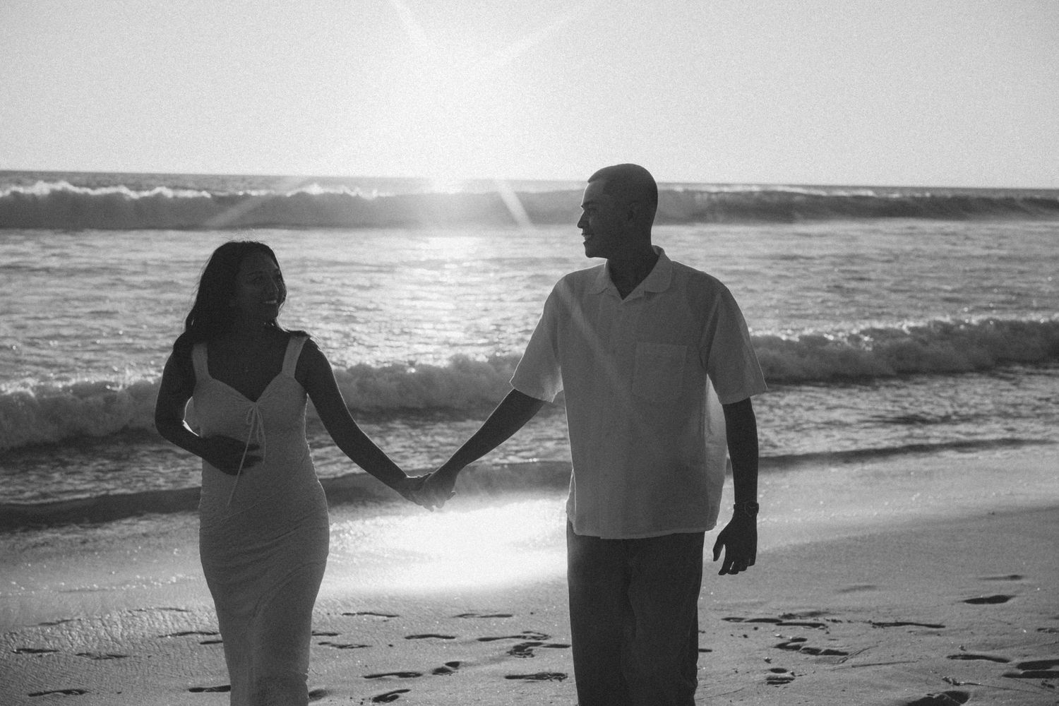Black and white photo of a couple holding hands walking on a sunny beach.