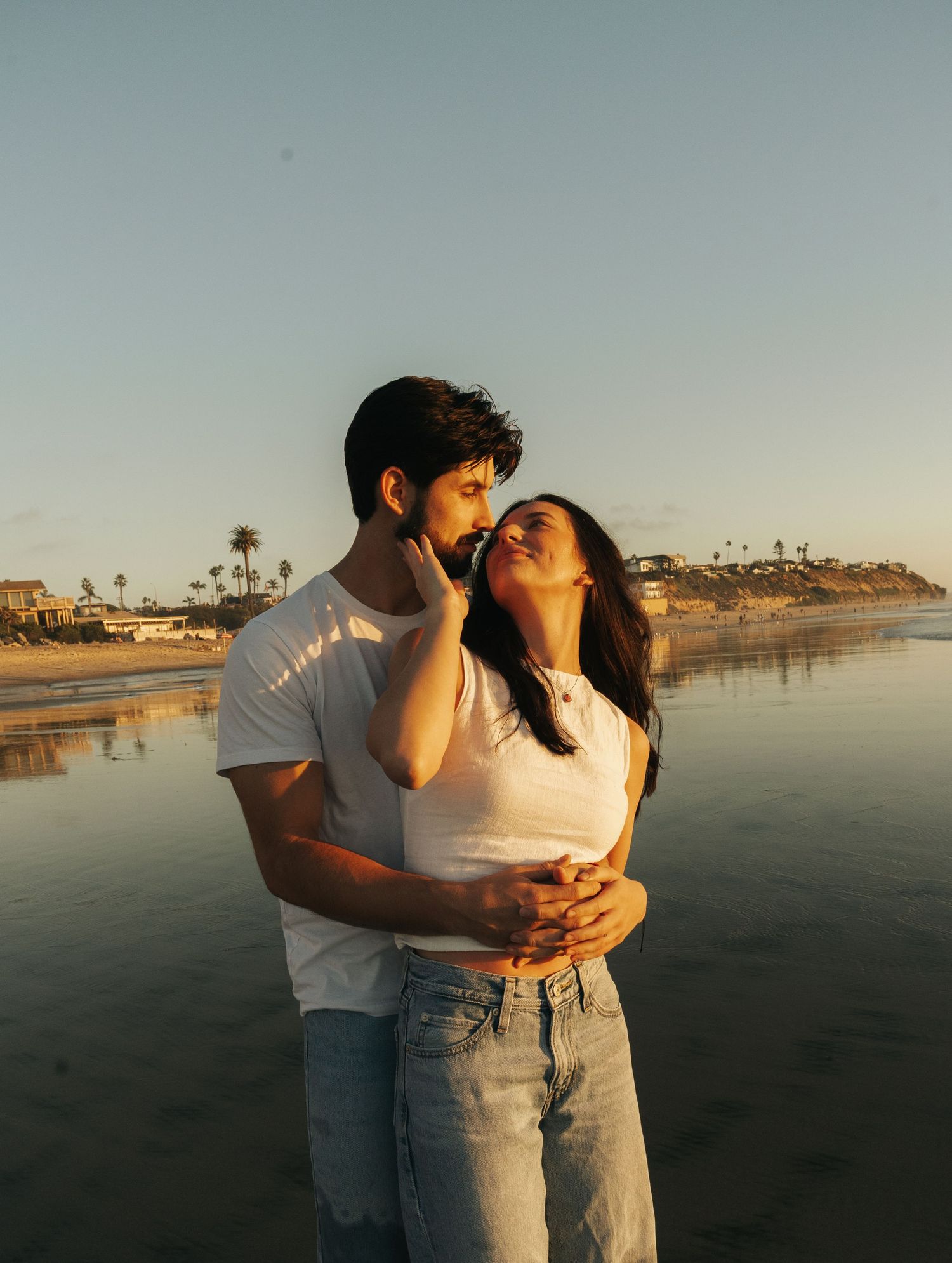 Romantic couple embracing on a San Diego beach at golden hour with ocean and cliffs in background.