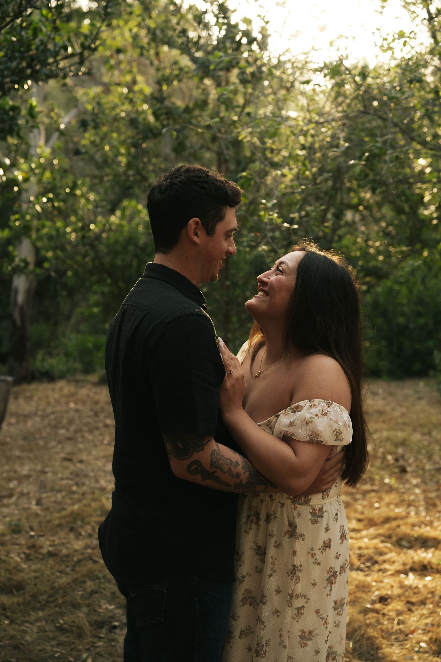 Couple gazing at each other and laughing while embracing in a sunlit forest setting.