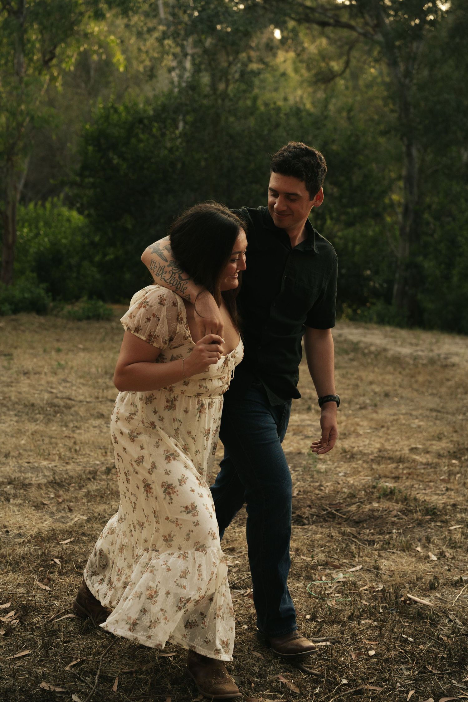 Couple dancing outdoors in a sunlit forest clearing, woman in floral dress, man in black shirt.