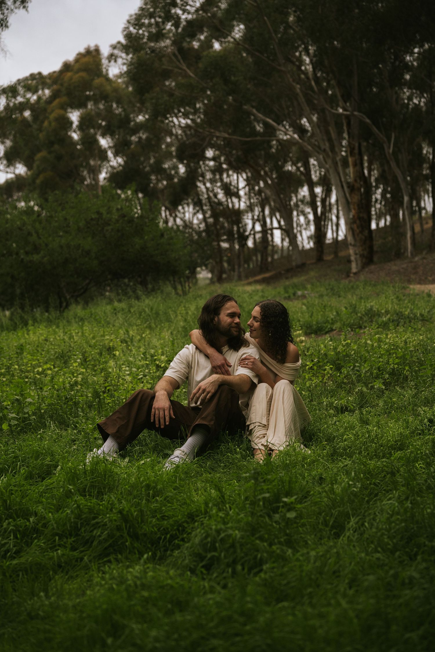 A couple sits together on lush green grass surrounded by trees, sharing a romantic moment in a serene outdoor setting.