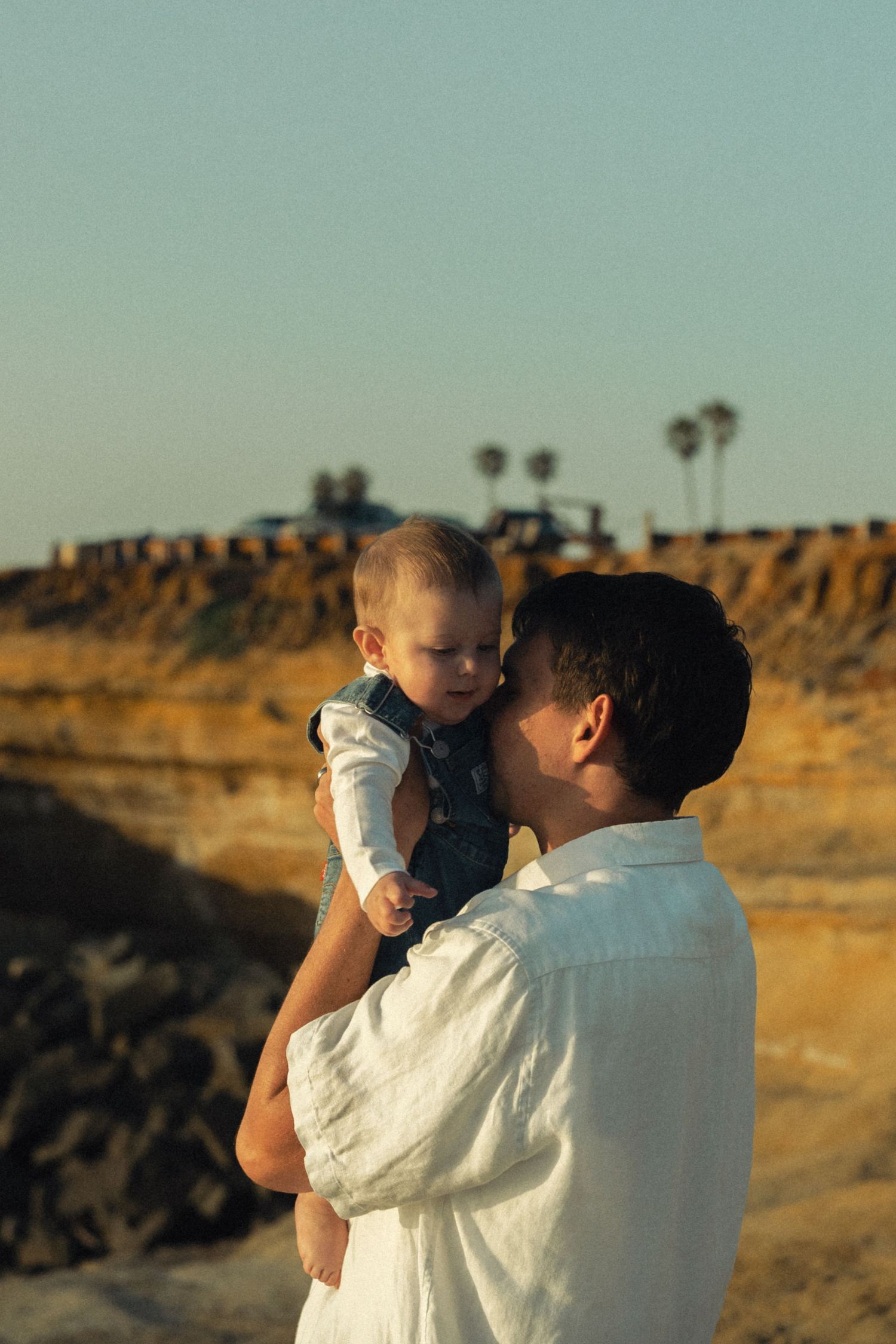 A father tenderly holds his baby boy near rocky cliffs with palm trees in the background at sunset.