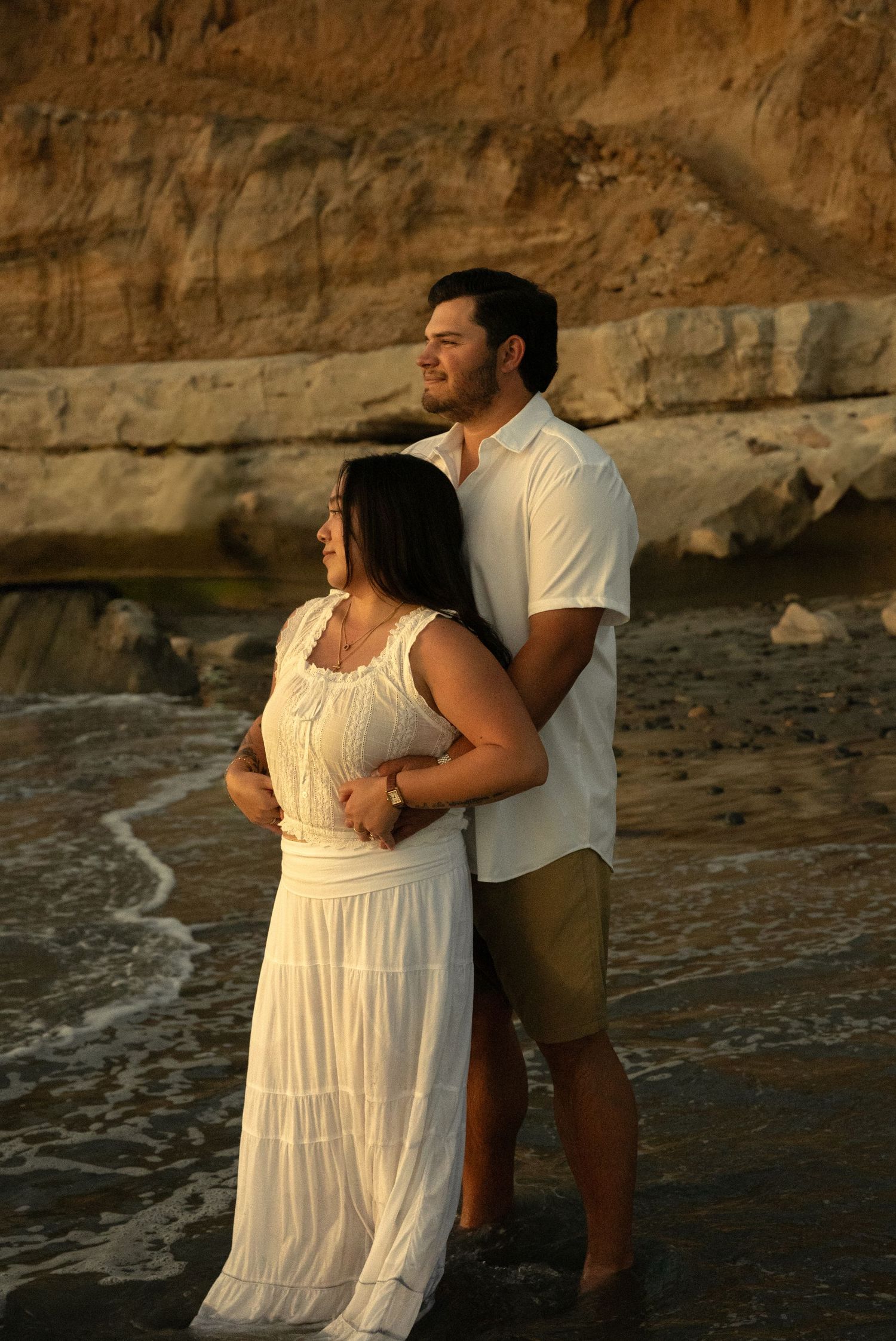 Couple embracing on rocky beach at golden sunset, woman in white dress, man in white shirt and khaki shorts.