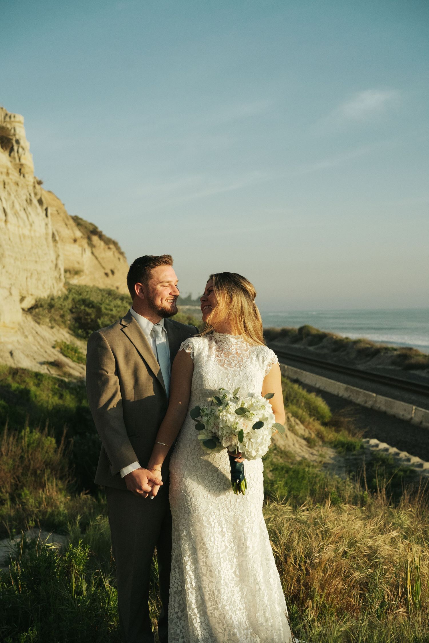 Smiling groom in tan suit holds brides hand near coastal cliffs with ocean backdrop at sunset.