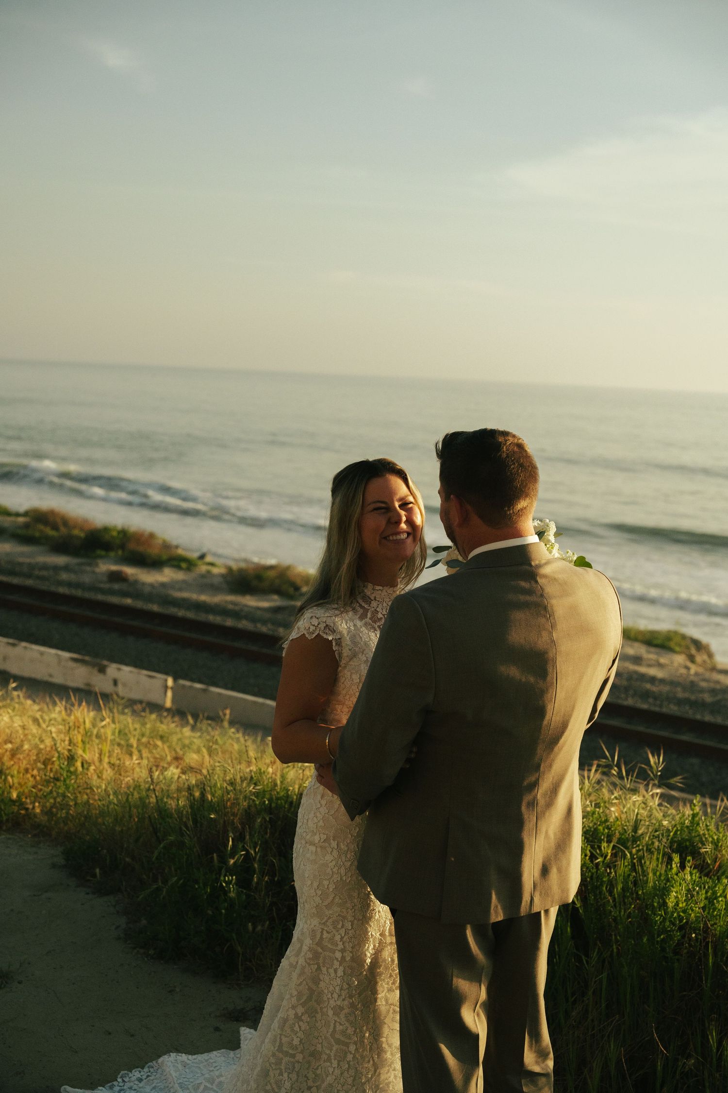 Bride and groom share a romantic moment by the ocean at golden hour, with waves and blue sky behind them.