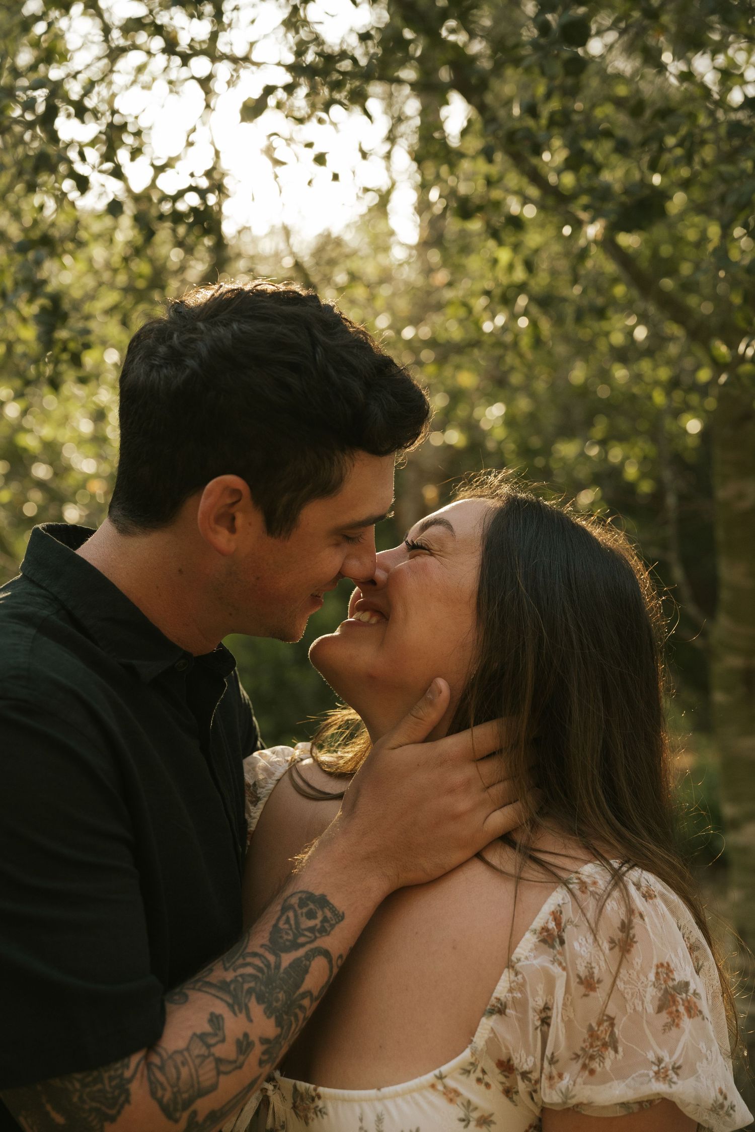 Couple sharing a tender almost-kiss in a sunlit forest, man with tattoos gently holding woman's face.