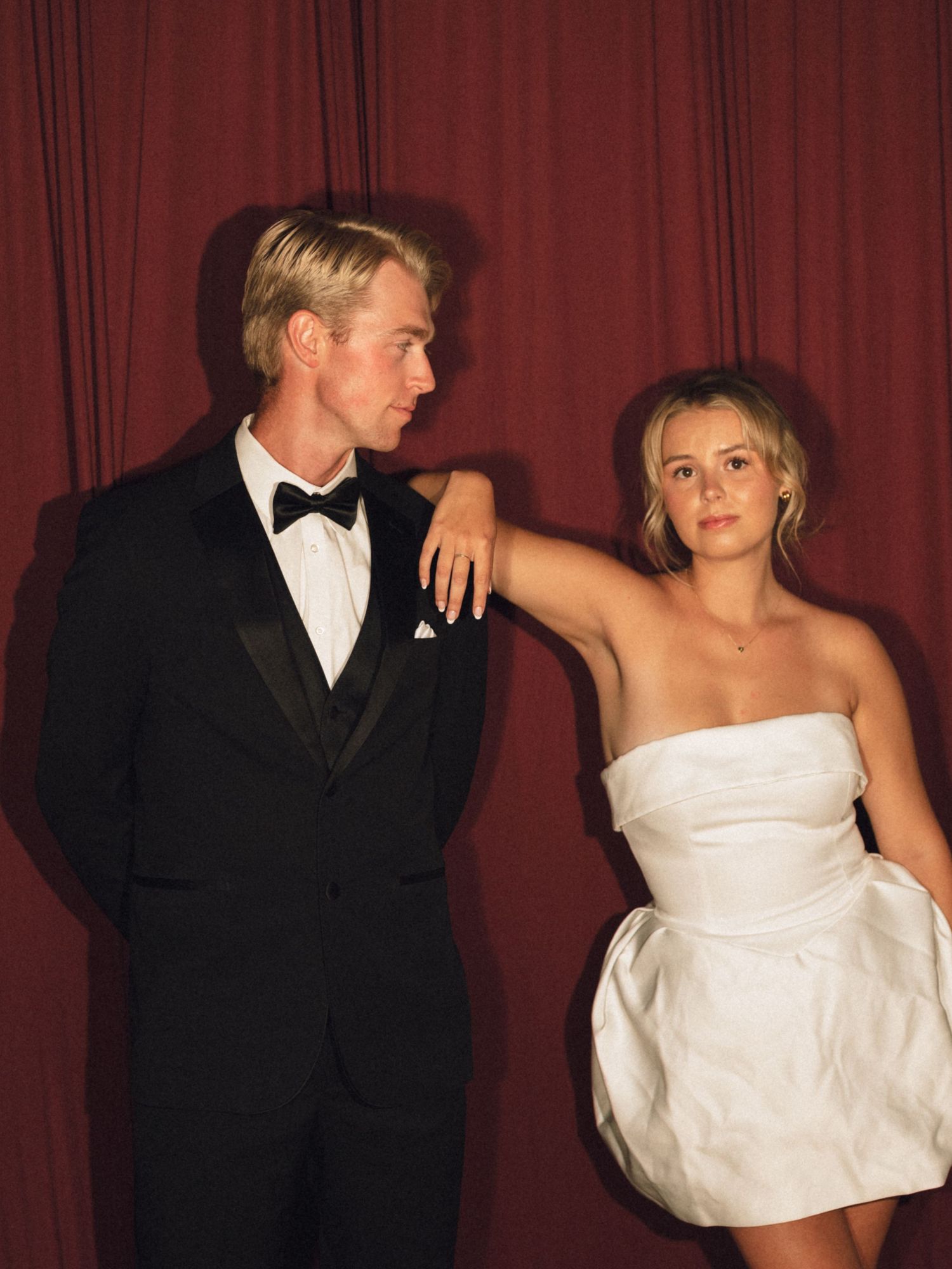 A man in a black tuxedo and a woman in a white strapless mini dress pose elegantly against a red curtain backdrop.