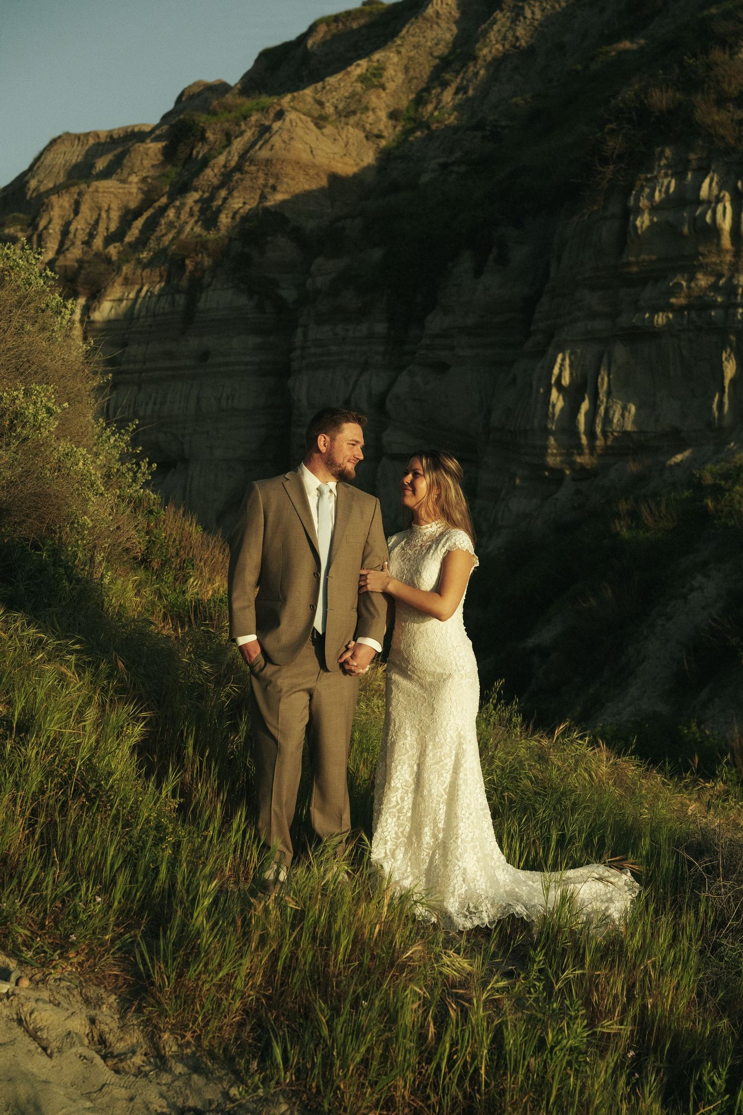 Bride and groom share a joyful moment on a grassy hillside with dramatic rocky cliffs in the background.