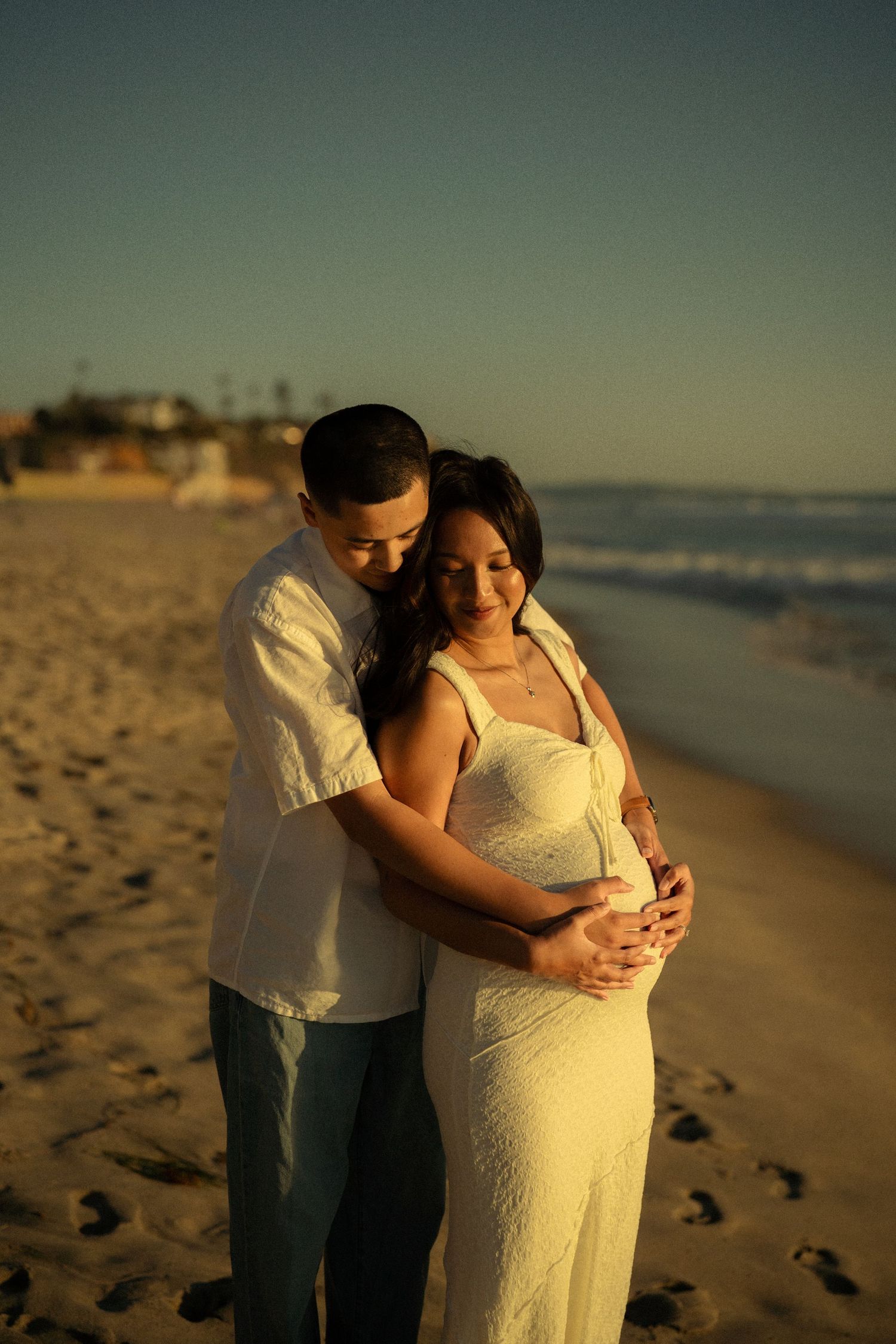 Couple embracing on beach at golden sunset, pregnant woman in white dress cradling baby bump.