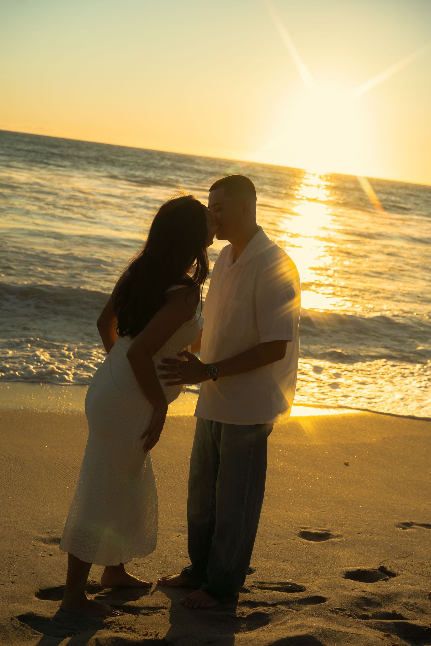 Pregnant couple sharing romantic kiss silhouetted against glowing golden beach sunset.